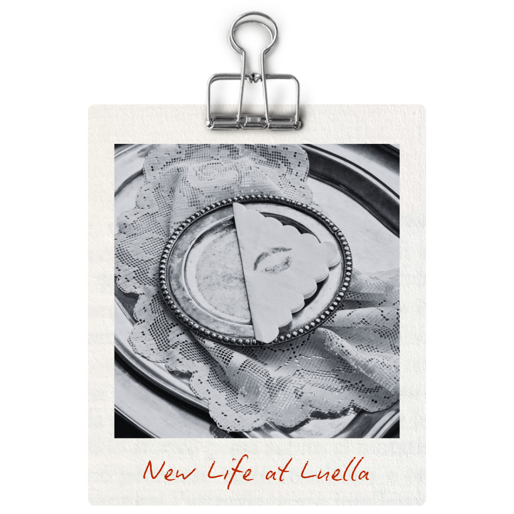 A silver tray with a lace cloth holding a divided scalloped-edge plate featuring a cake with a design of an eye. The photo is black and white with the caption "New Life at Luella" written in red at the bottom.