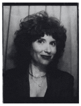 Black and white photo of a woman with curly hair smiling, wearing a blazer and a necklace, standing in front of a paneled wall.