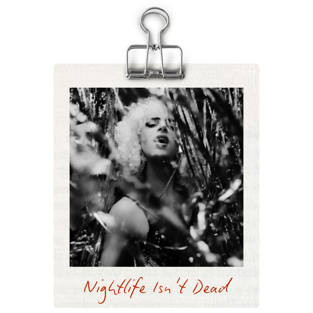 A black and white photograph of a woman with curly hair among tall plants or grass, her eyes closed and mouth open, suggesting a sensual or emotional expression. The photo is held on a piece of paper clipped with a metal binder clip, and the text 'Nightlife Isn't Dead' is handwritten in red at the bottom of the paper.