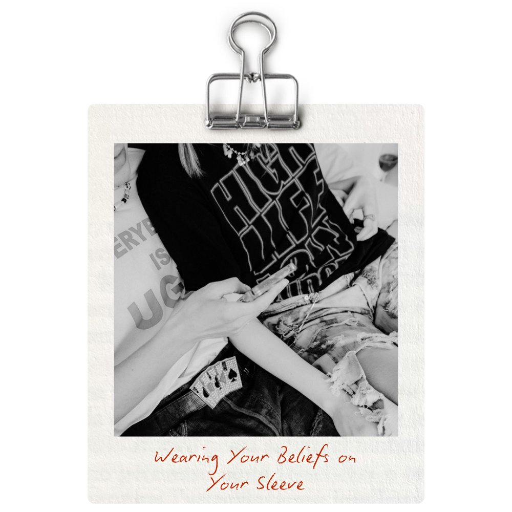 A black and white photo of a person sitting, wearing a black T-shirt with logo text, ripped denim shorts, and accessories, holding a smartphone. The photo is clipped to a piece of paper with handwritten text in red ink that says, "Wearing Your Beliefs on Your Sleeve."