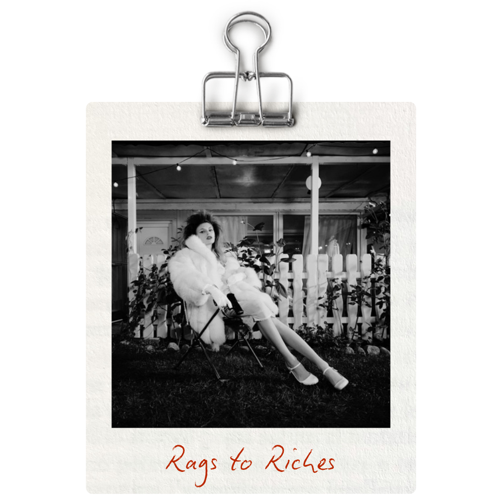 Black and white photo of a woman sitting on a chair outside, wearing a fur coat and heels, with a white picket fence and string lights in the background. Text at the bottom reads "Rags to Riches."