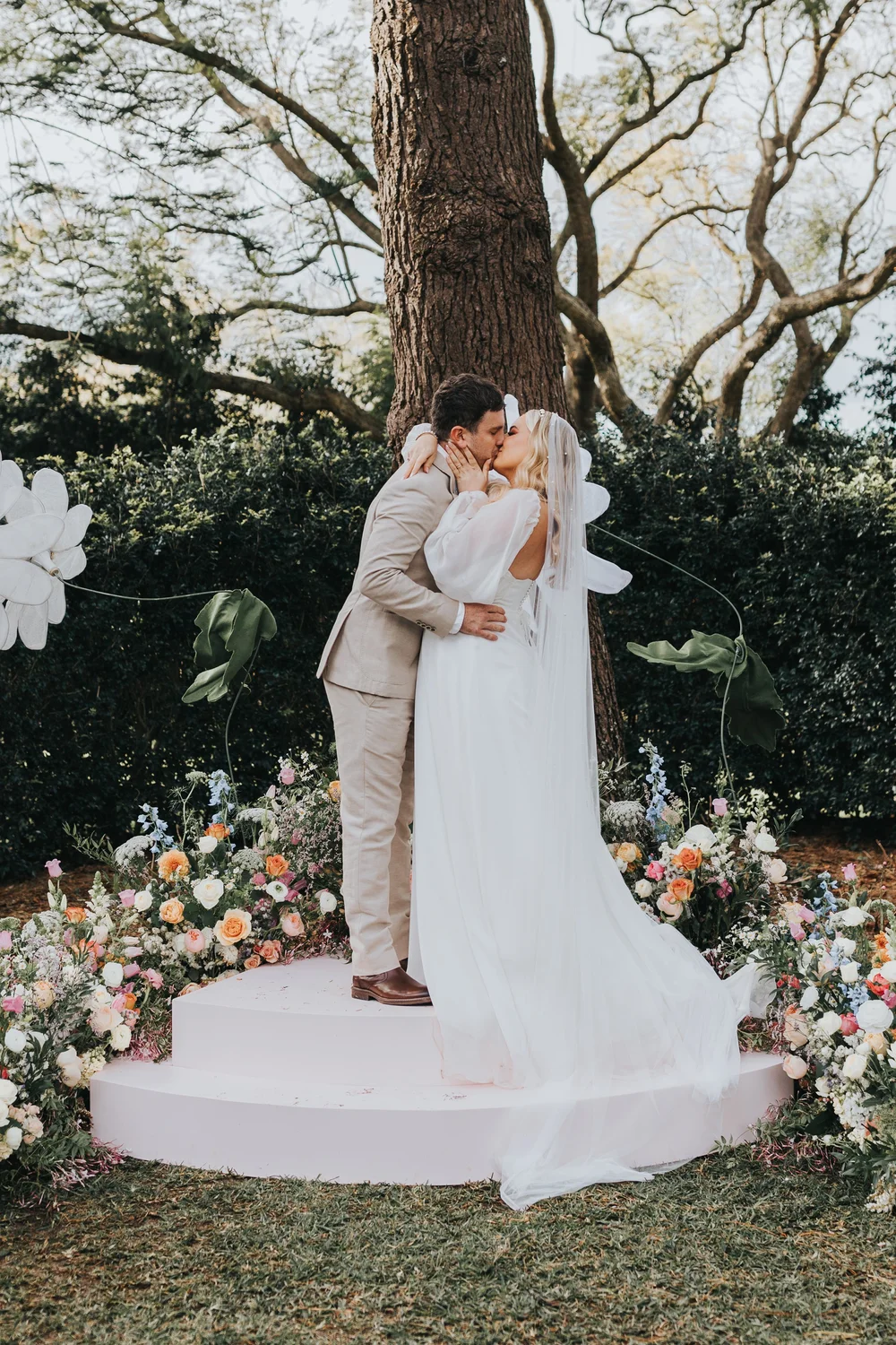 Bride and groom kissing on a flower-covered stage under a tree