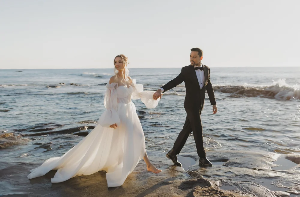 Bride and groom walking hand in hand across rocks by the sea