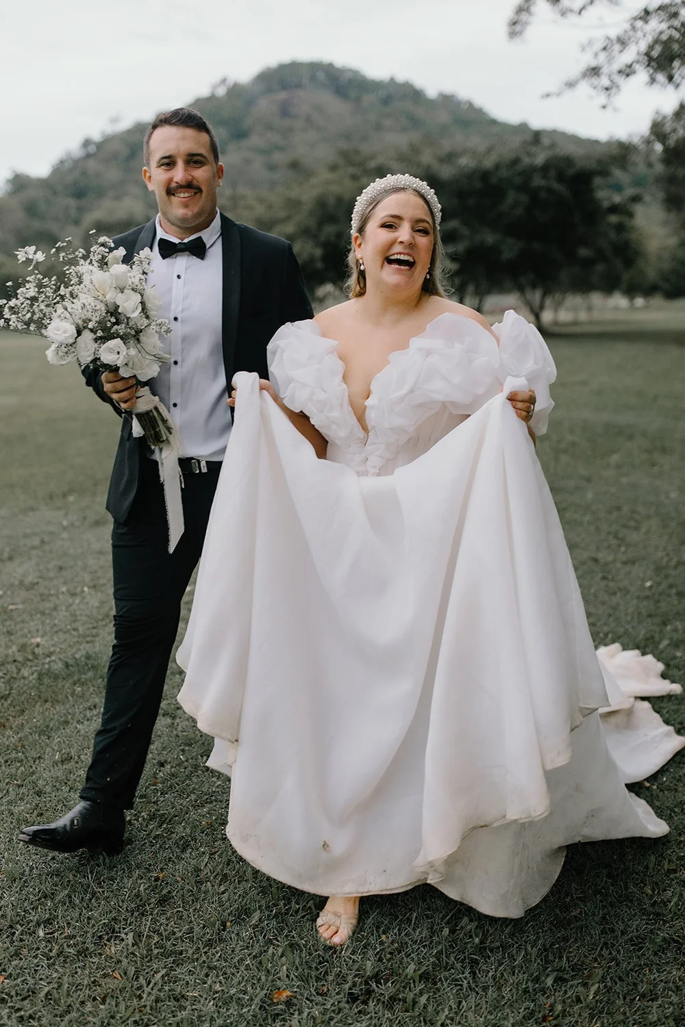Laughing bride lifting her skirt beside her groom, with mountains behind