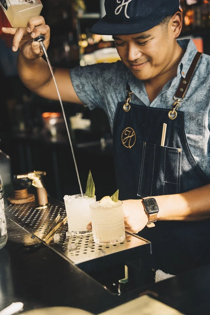 A bartender preparing cocktails at a bar, pouring liquid from a squeeze bottle into two glasses decorated with lime wedges and ice.