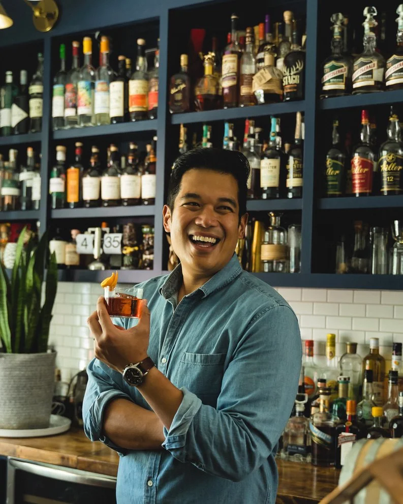 A man smiling and holding a cocktail glass in a bar with shelves filled with various liquor bottles behind him.