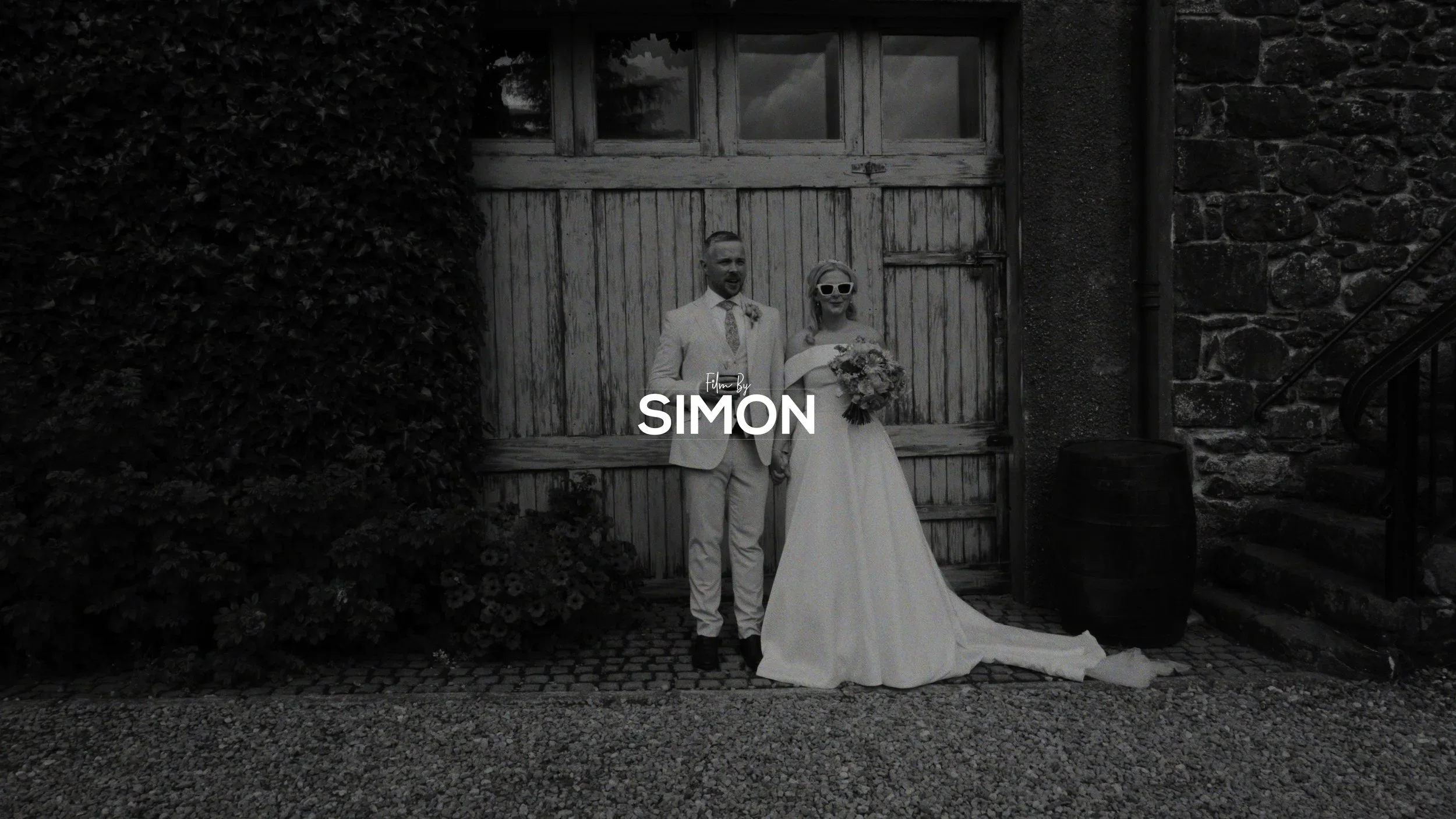 Black and white photo of a bride and groom standing outside in front of a wooden garage door. The groom is dressed in a light-colored suit and tie, and the bride in a wedding gown holding a bouquet. The bride is wearing sunglasses. There is a large bush and stone wall on either side, and a barrel near the stairs.
