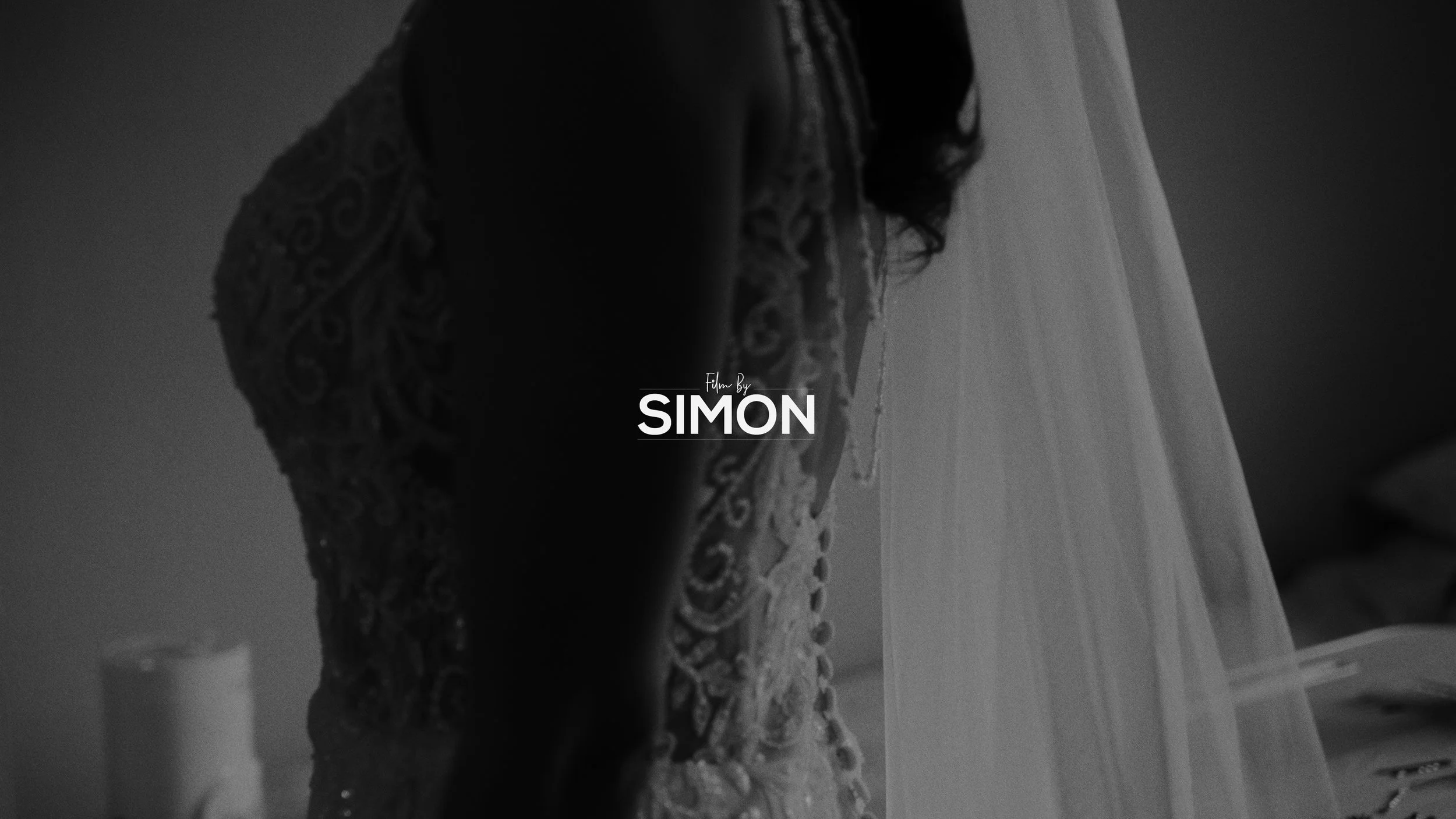 A woman in an intricate lace dress with a sheer veil over her head, standing indoors with a blurred background.