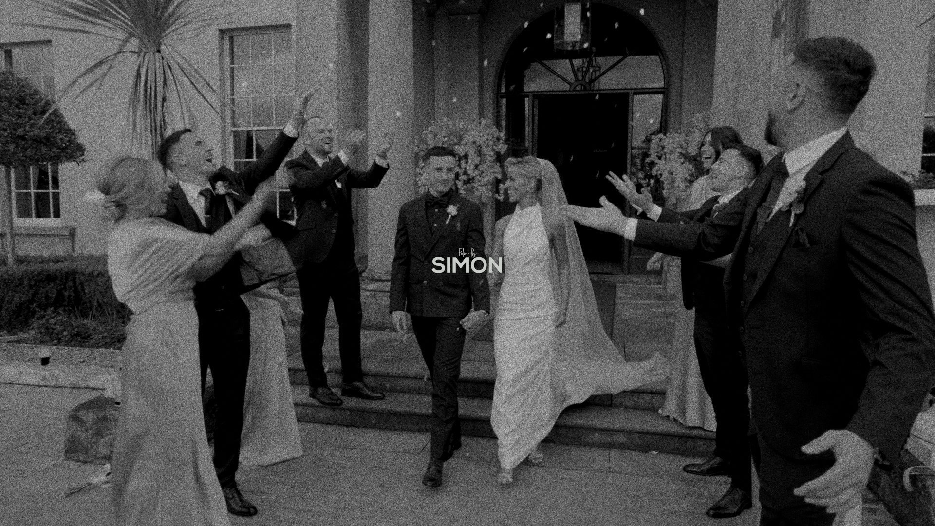 A black and white photo of a wedding celebration outside a building with stairs. The bride and groom are walking down the stairs hand in hand, smiling. Guests are on either side, cheering and reaching out their hands toward the couple.