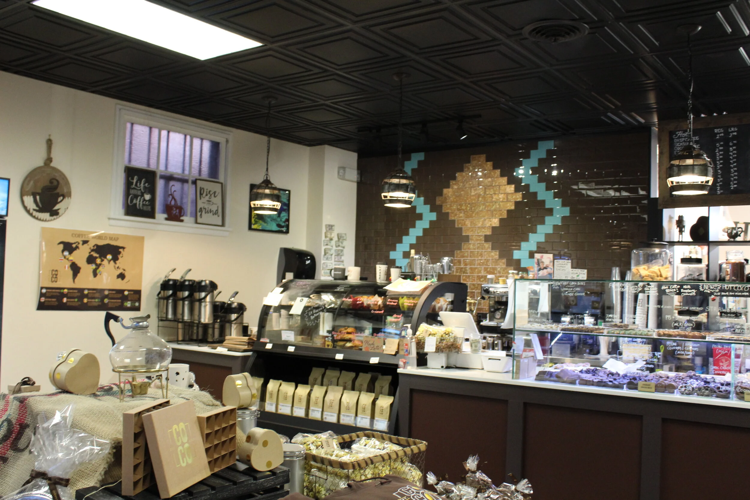 Interior of a coffee shop with a display counter of baked goods, coffee brewing equipment, decorative wall art, and a coffee-related theme on the wall.