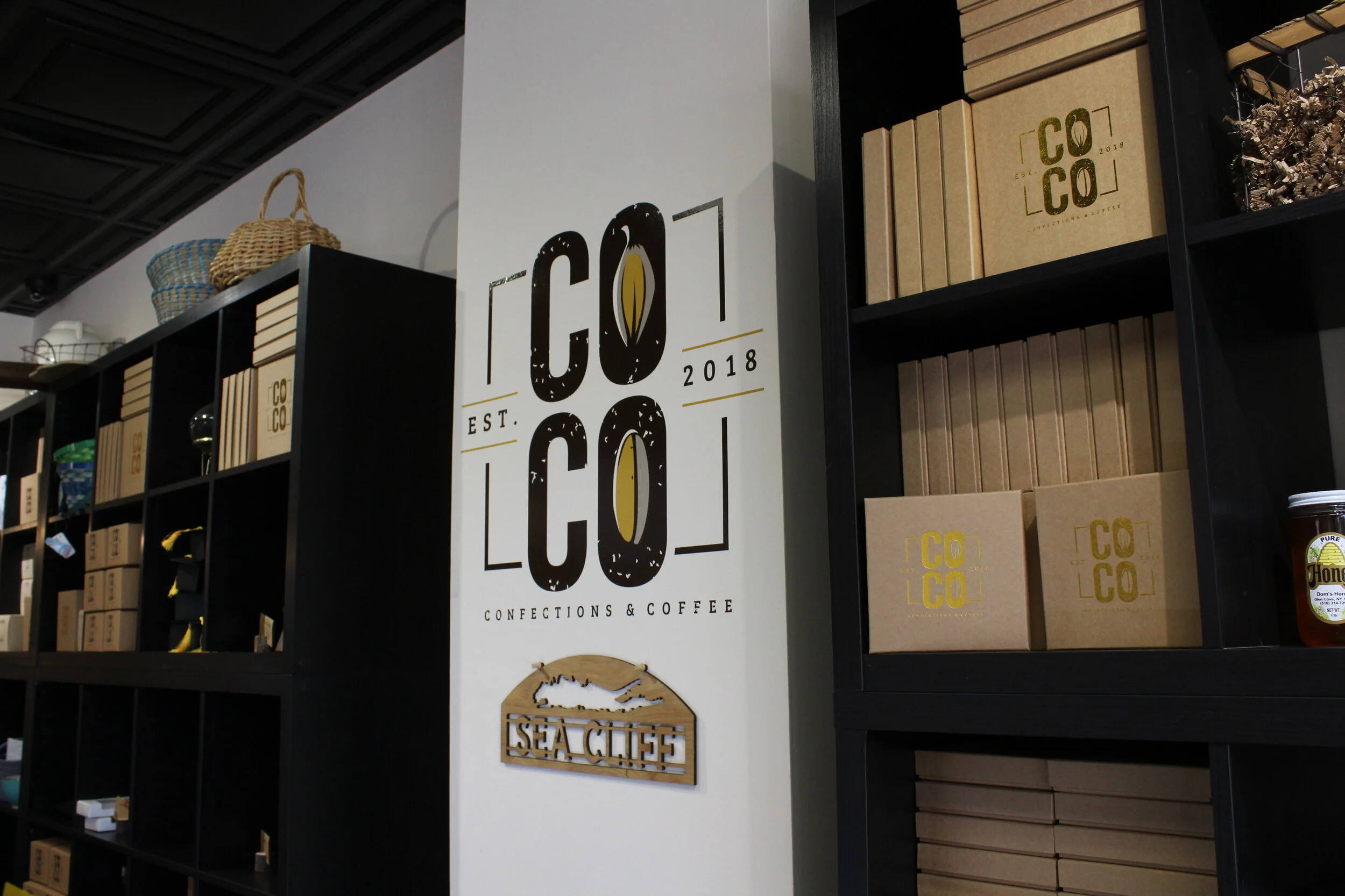 Interior of a cafe or store with black shelving holding boxes and baskets, and a large central sign with the logo 'COCO' and phrases 'CONFECTIONS & COFFEE' and 'EST. 2018', featuring a coffee bean and the words 'SEA CATCH' with a fish silhouette.