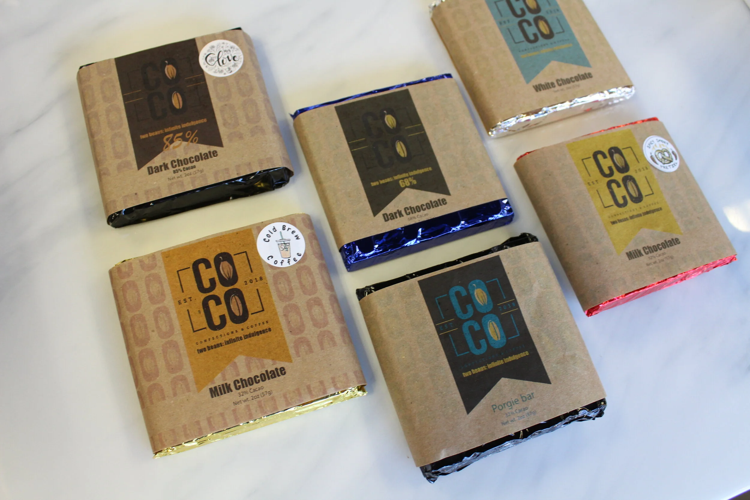 Six bars of chocolate and a coffee bar on a white surface, each wrapped in brown paper with labels indicating flavors like dark chocolate, milk chocolate, white chocolate, and porgie bar.