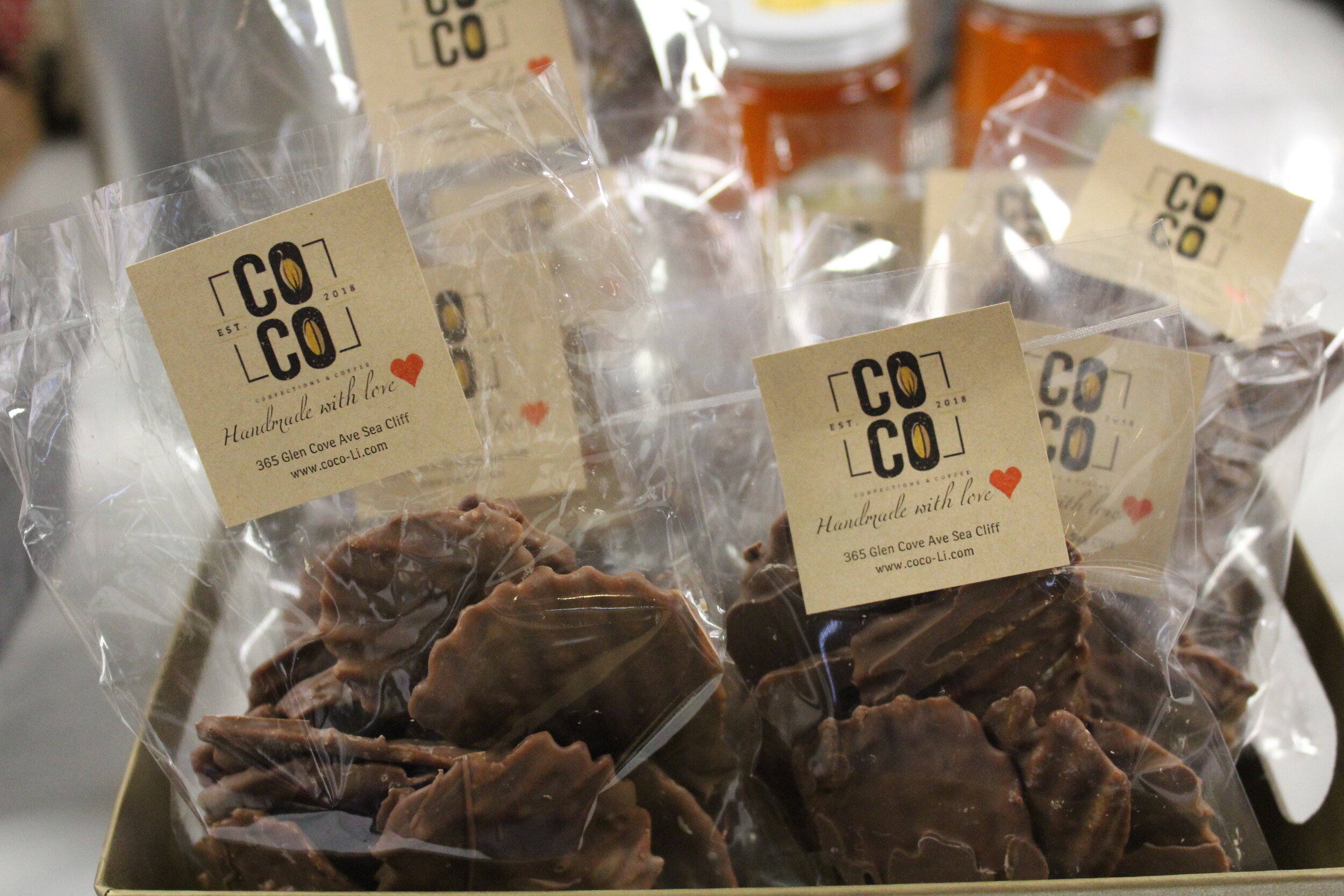 Bags of handmade chocolate treats with labels from Coco, featuring the address 365 Glen Cove Ave Sea Cliff, with jars of honey in the background.