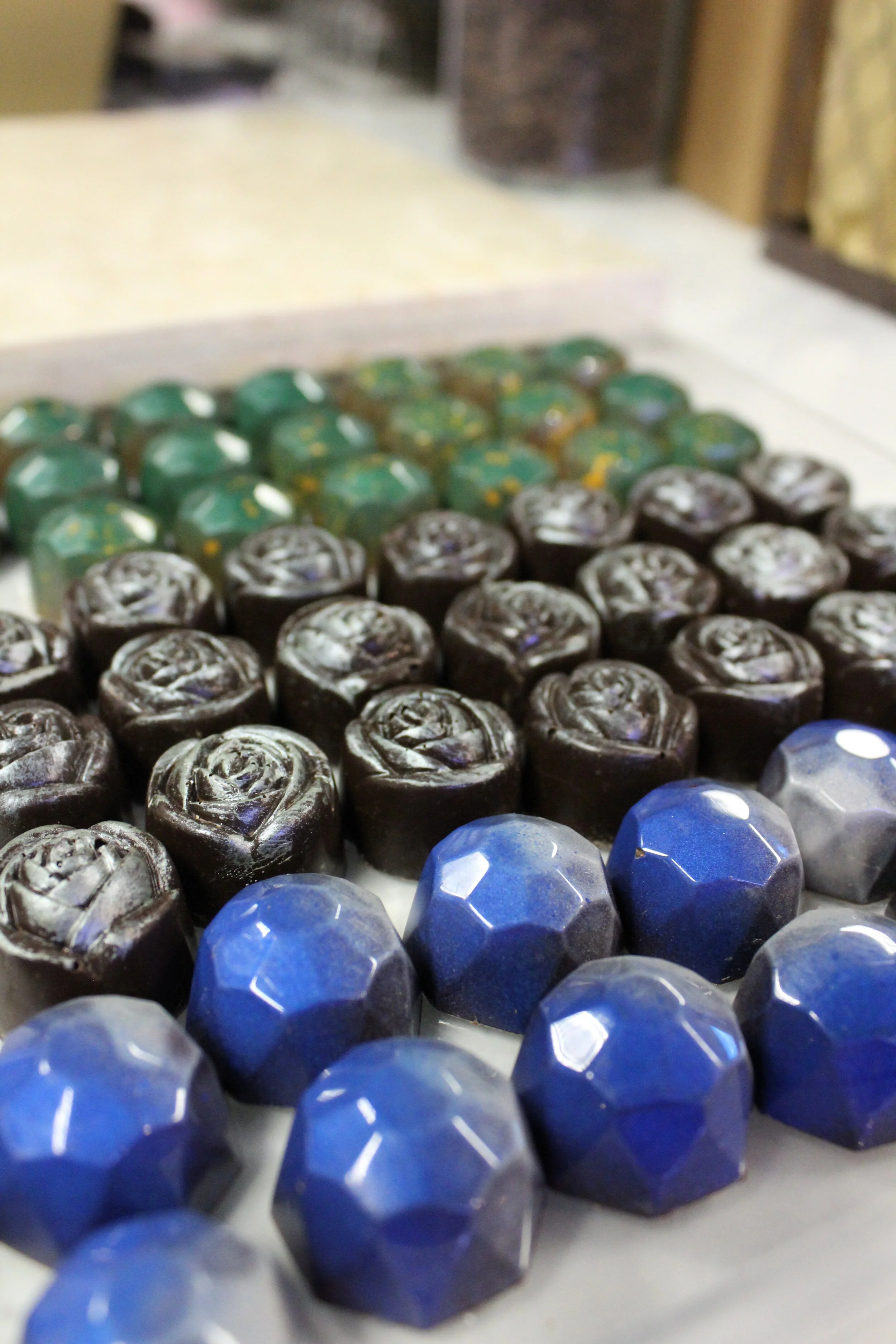 Assorted chocolates arranged in rows, including green, black, and blue pieces with various shapes and patterns, on a tray.