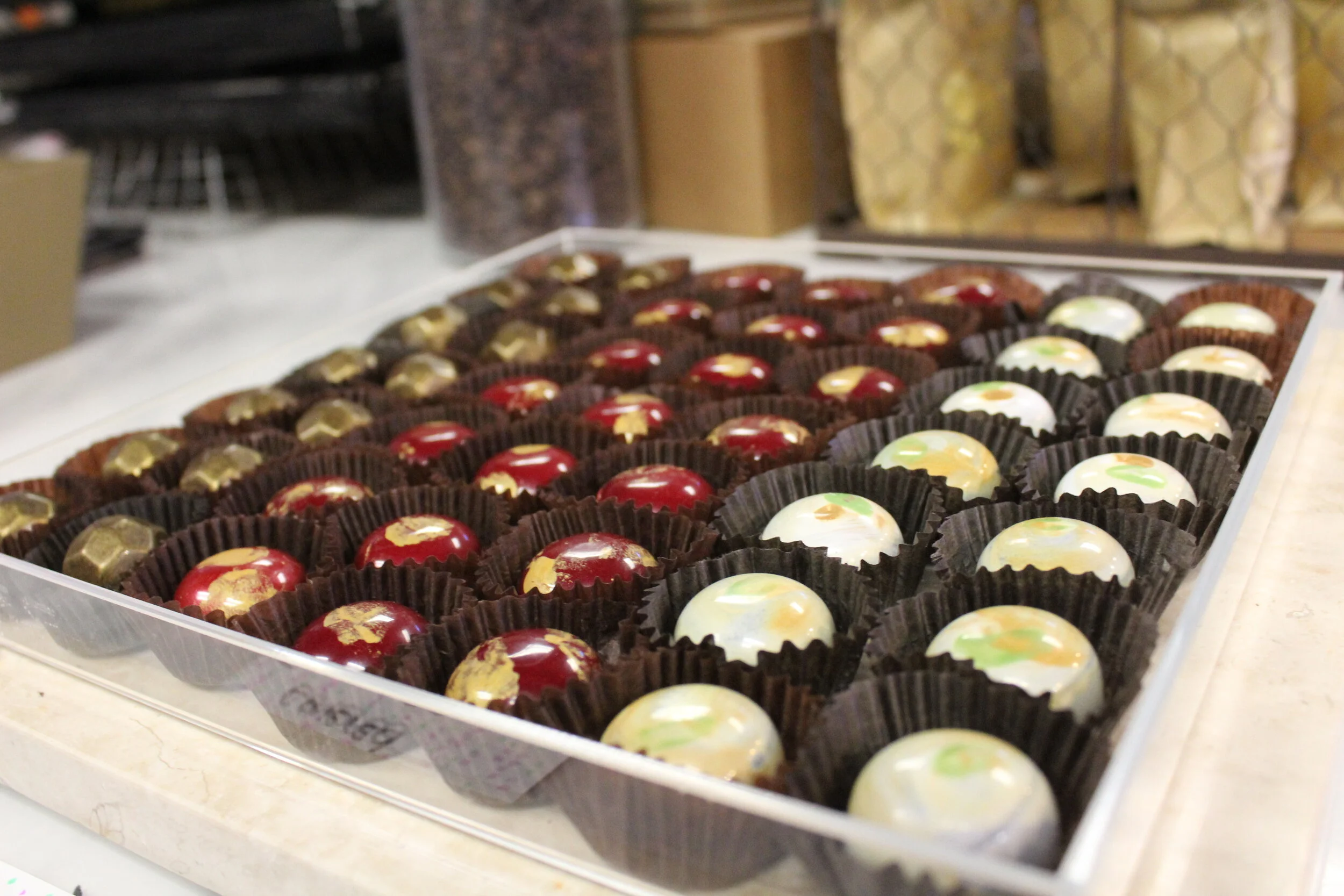 A tray of assorted chocolates in black paper cups with white, red, and brown coatings decorated with gold and green accents.