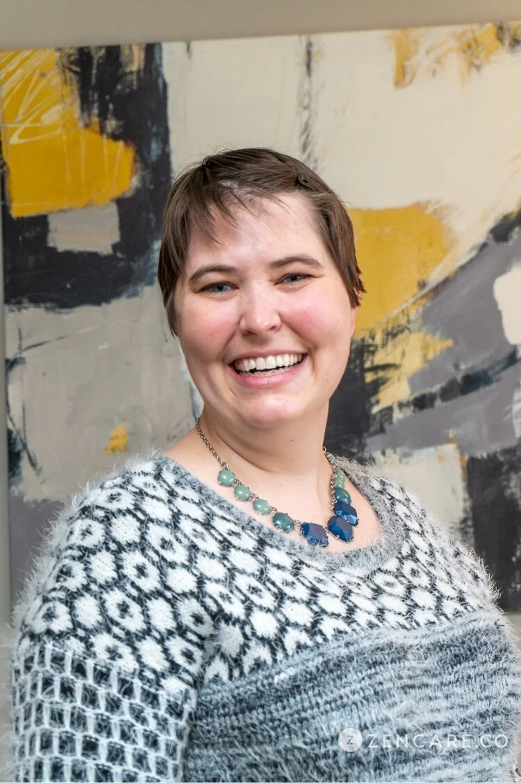 A smiling woman with short brown hair wearing a patterned sweater and a blue necklace, standing in front of an abstract art background.