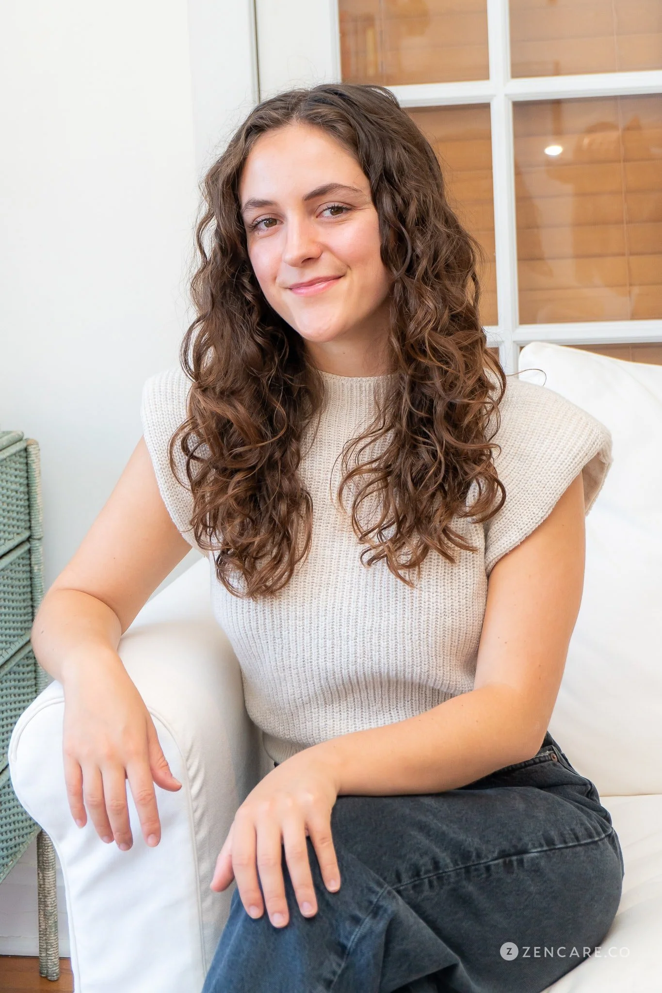 A young woman with curly brown hair sitting on a white couch, smiling at the camera.