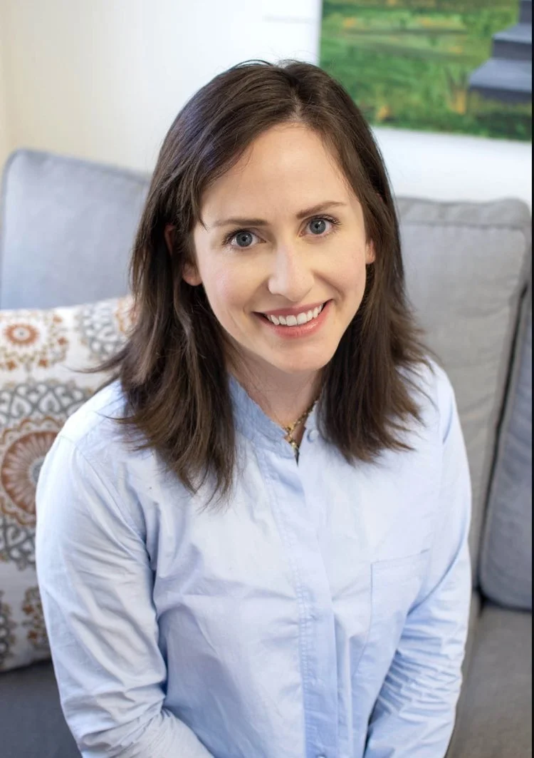 A woman with shoulder-length brown hair and blue eyes smiling at the camera, sitting on a light-colored couch in a living room.