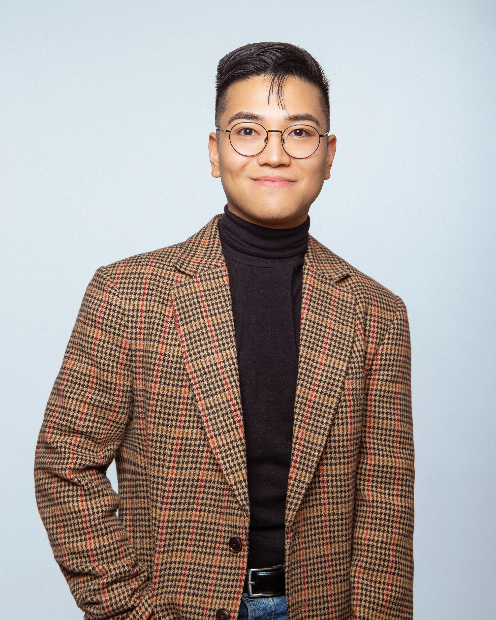 Young man with short black hair, glasses, wearing a brown checked blazer, black turtleneck, and blue jeans, smiling at the camera against a light gray background.