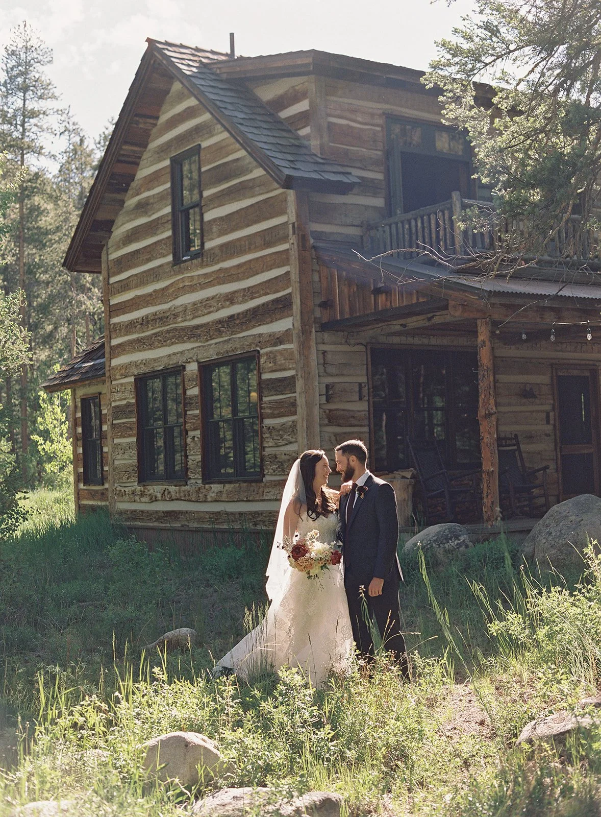 Bride and groom in wedding attire standing outside cabin in the sun embracing