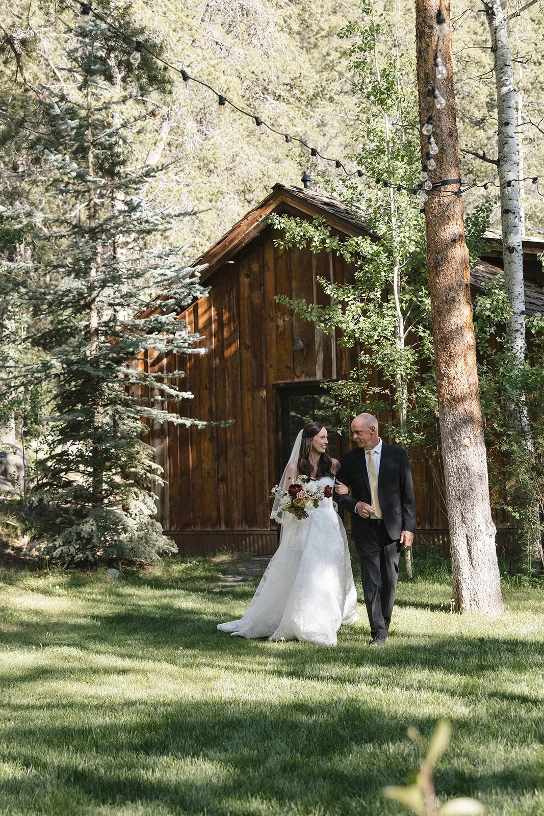 Bride walking with her dad in a tux in field with cabin behind them