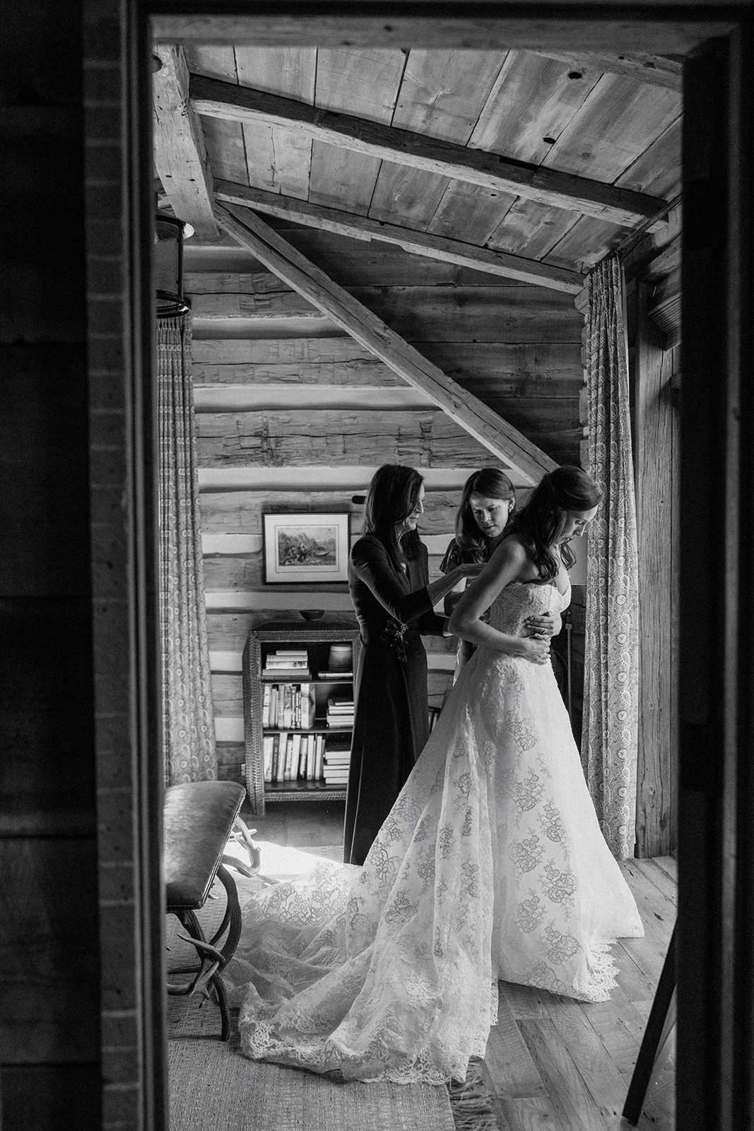 Bride putting on wedding dress with mom and sister buttoning up back inside of cabin