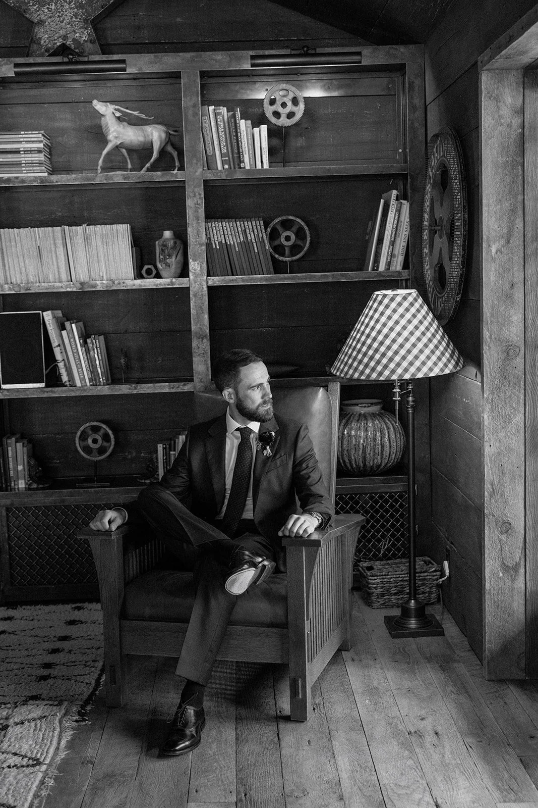 Groom sitting in leather arm chair in cabin with bookshelves behind looking out window