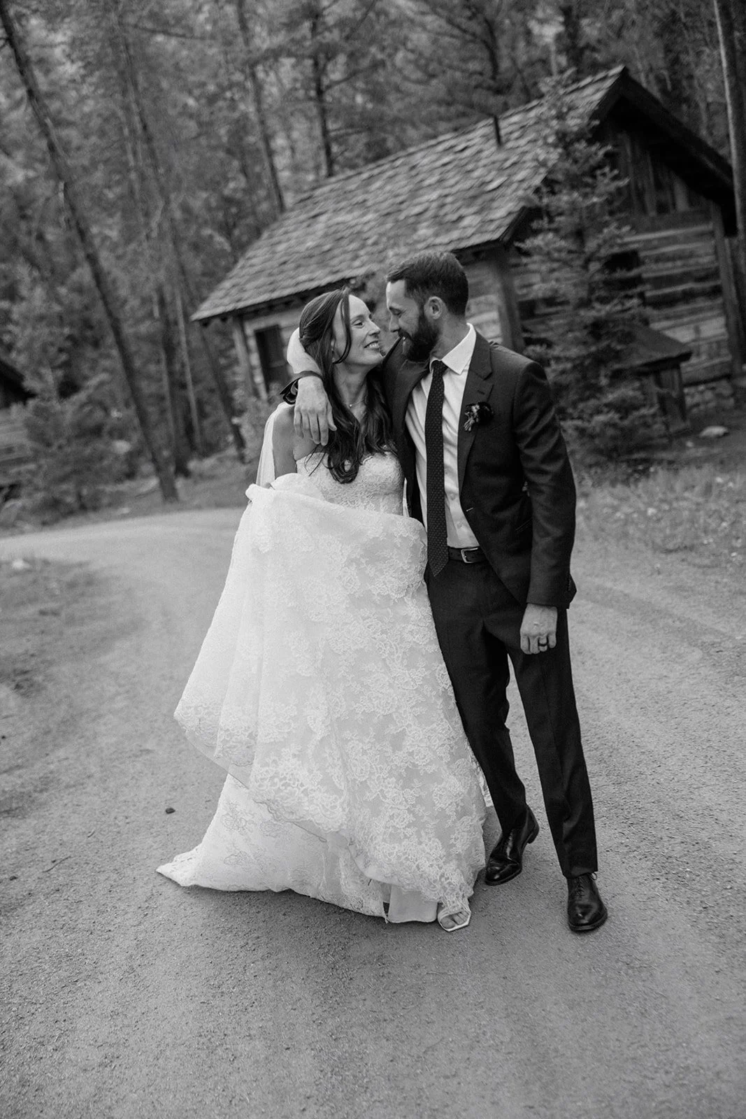 Bride and groom walking along path with cabins behind embracing in wedding attire