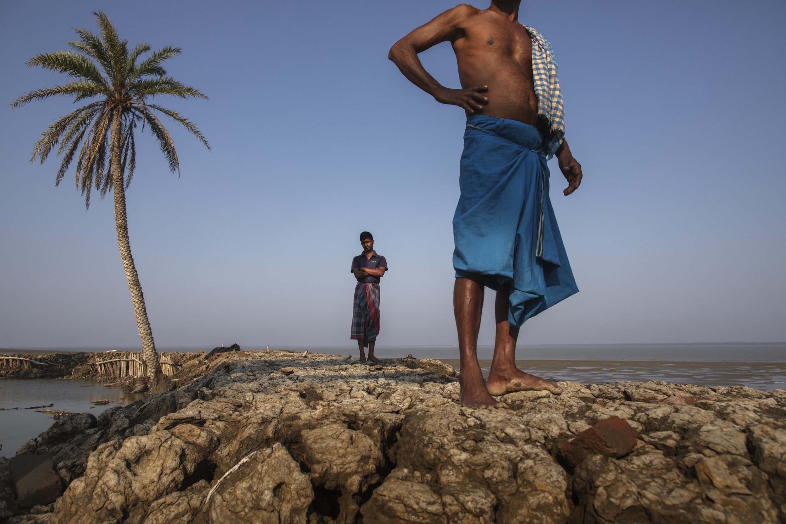  Saheb Dalui and Subhodip Bhuinya stand on a man-made seawall, mainly made out of soil and bamboo. The barrier protects their houses and livelihood getting submerged by the rising sea levels, Sundarbans/ India - 2019 