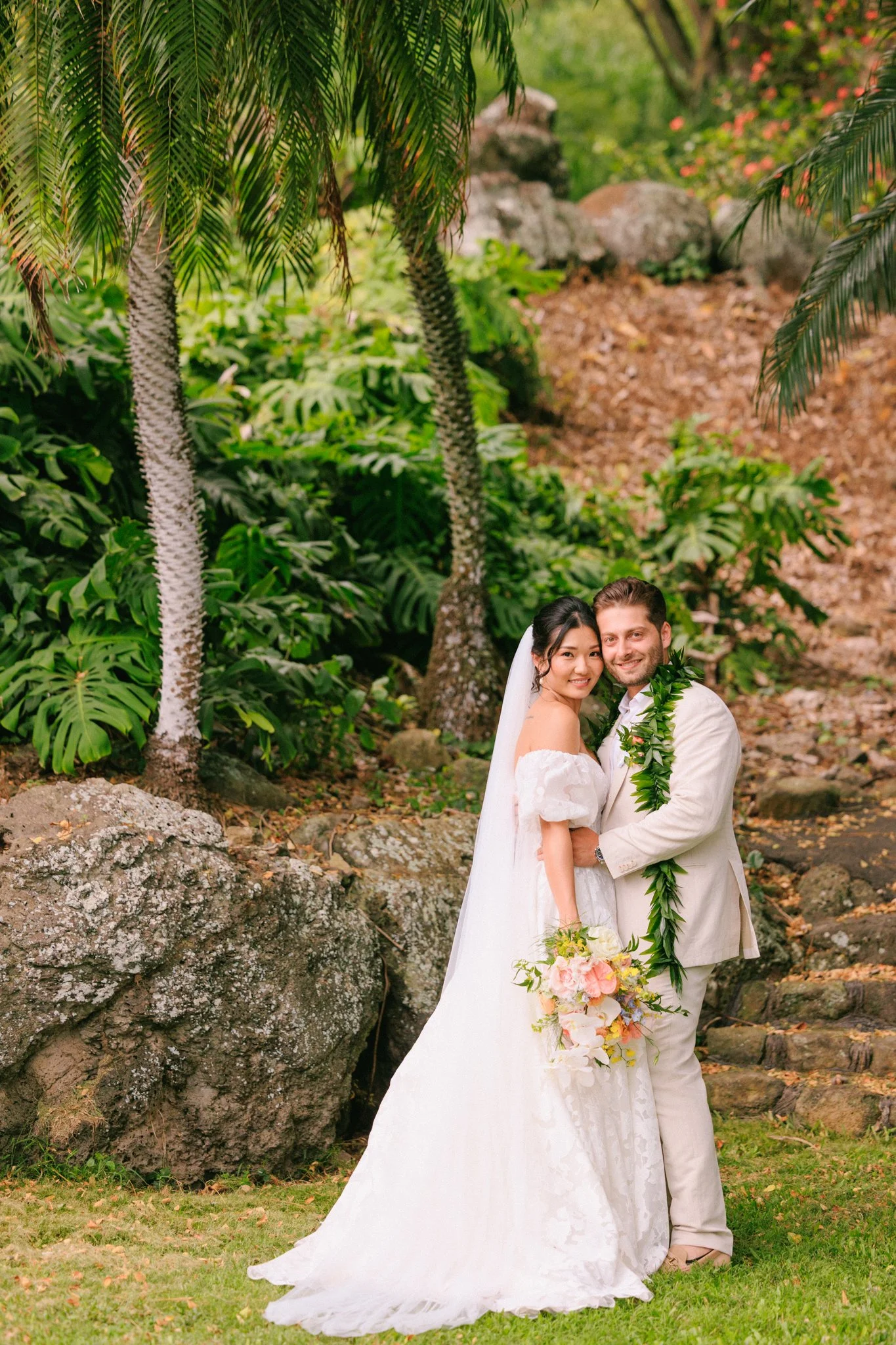 A bride and groom in wedding attire smiling and embracing outdoors in a lush, tropical garden with large rocks, green foliage, and palm trees.