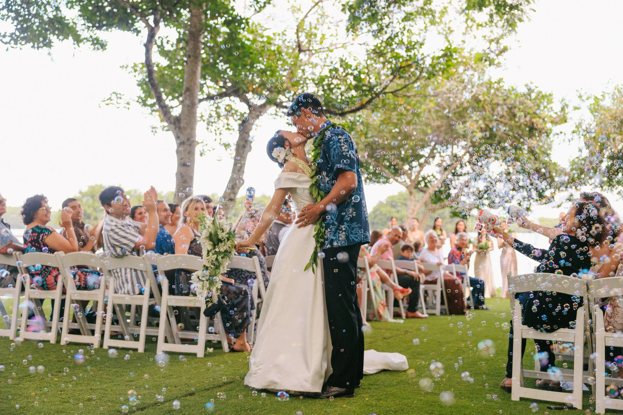 A newlywed couple sharing a kiss at their outdoor wedding ceremony, surrounded by seated guests and trees, amid bubbles in the air.