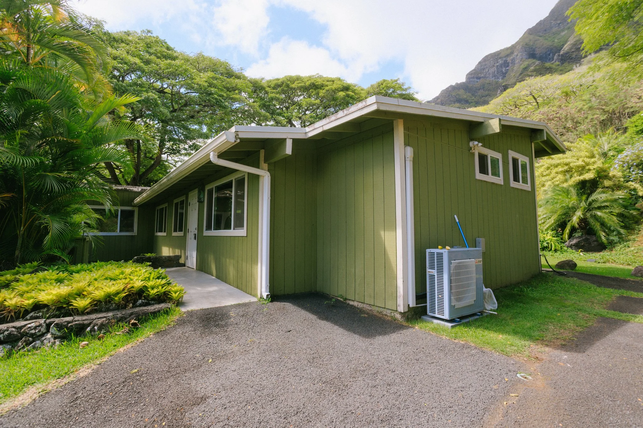 A green house with white windows and trim, surrounded by lush greenery and trees, with mountains in the background and a bright blue sky.
