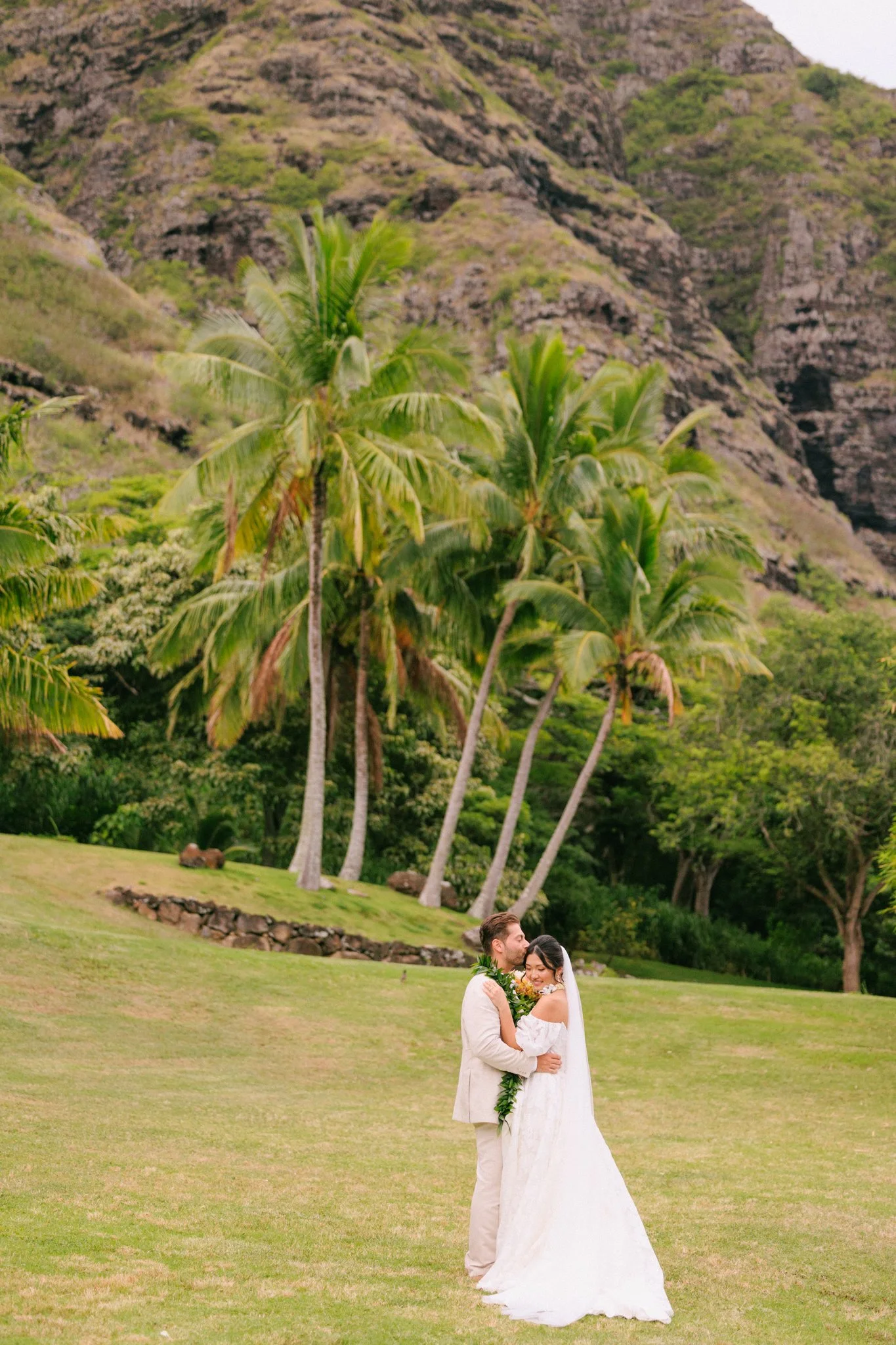 A bride and groom embracing outdoors on a grassy field with palm trees and mountain scenery in the background.