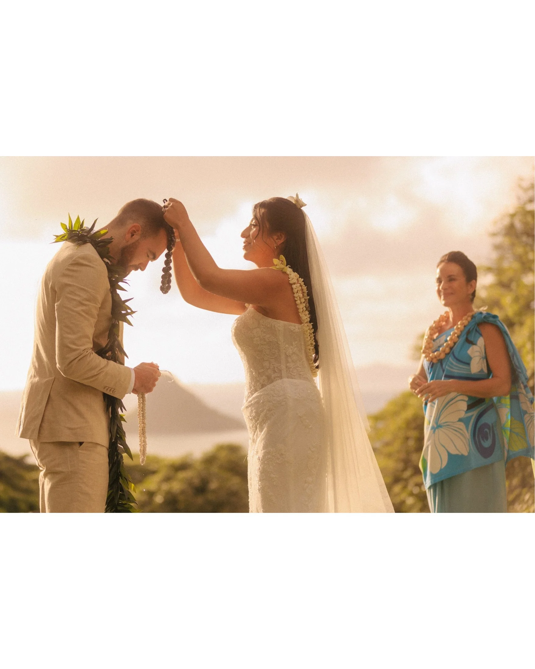 Nicole places a lei over Ethan's bowed head during their sunrise ceremony at Paliku Gardens, Kualoa Ranch, while their officiant watches from the side in warm morning light on Oahu, Hawaii.