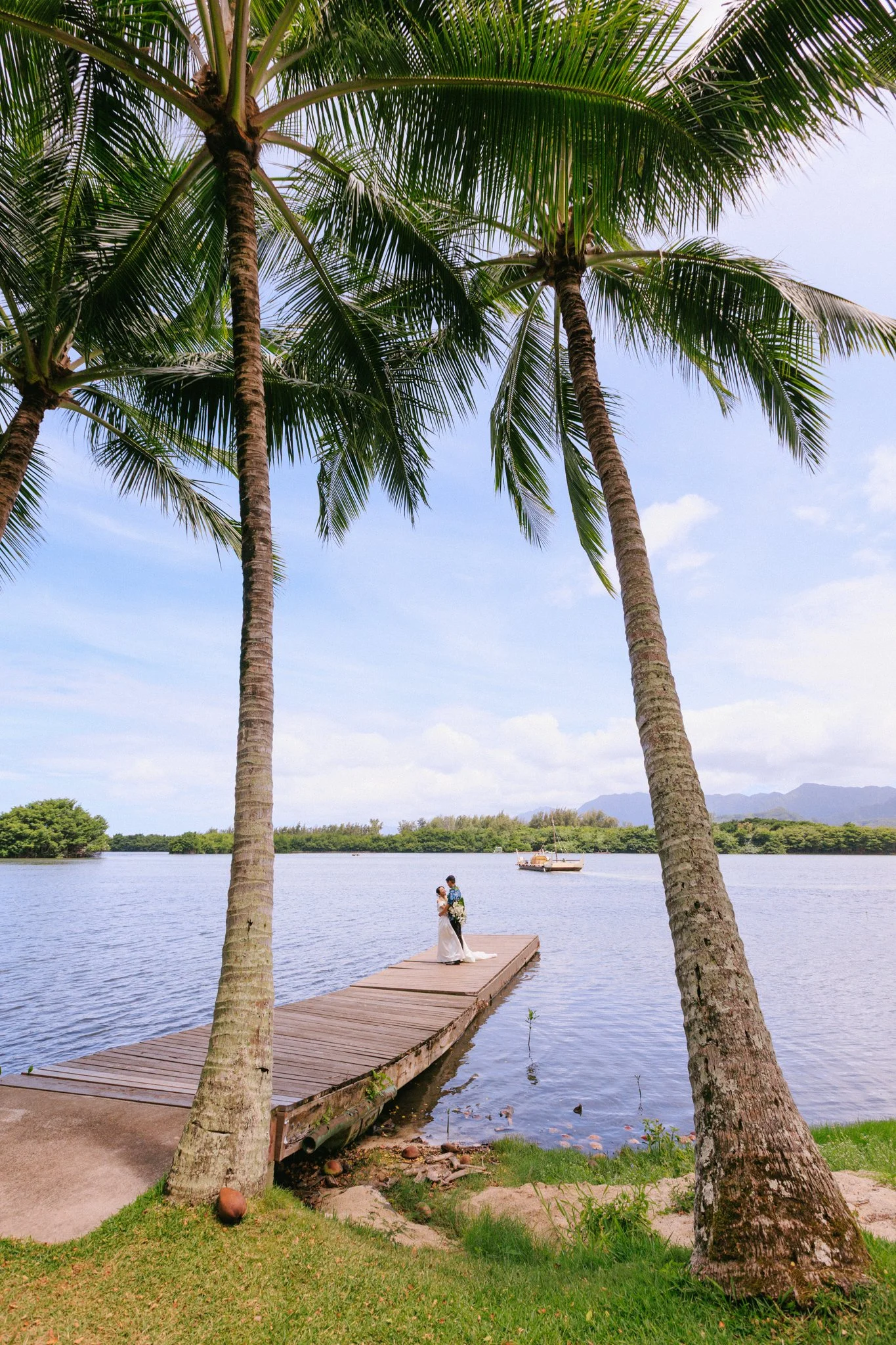 A couple in wedding attire standing on a wooden dock by a lake, surrounded by palm trees under a blue sky.