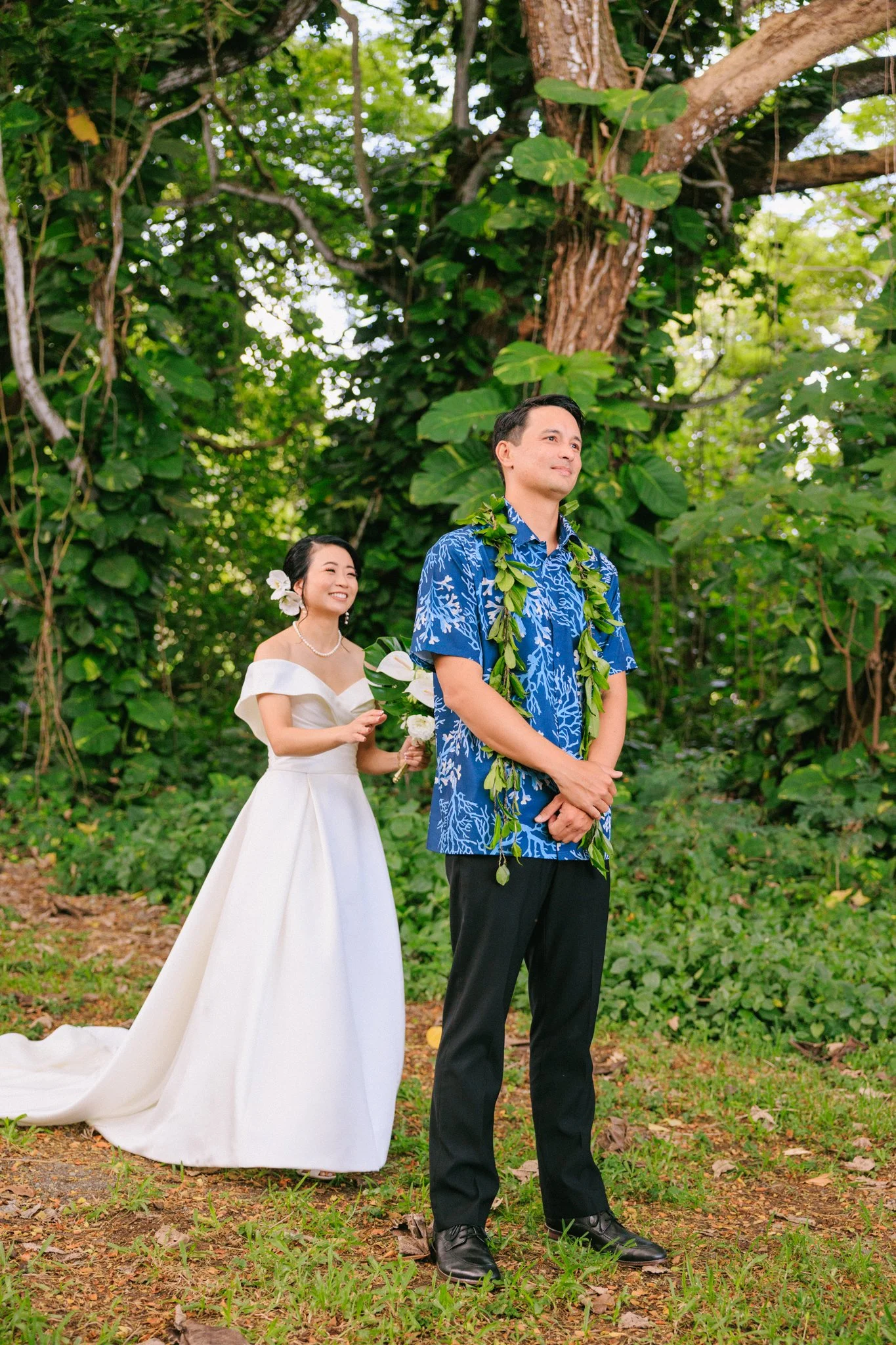 A bride and groom standing outdoors in a lush green forest, with the bride smiling and holding flowers, and the groom wearing a blue Hawaiian shirt with a lei, both looking content.