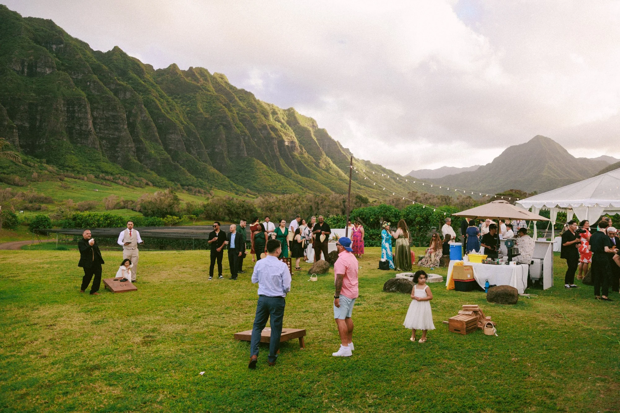 Guests gathered at an outdoor event in a lush green field with mountainous landscape in the background, some standing, some sitting, and kids playing, under tents and string lights.