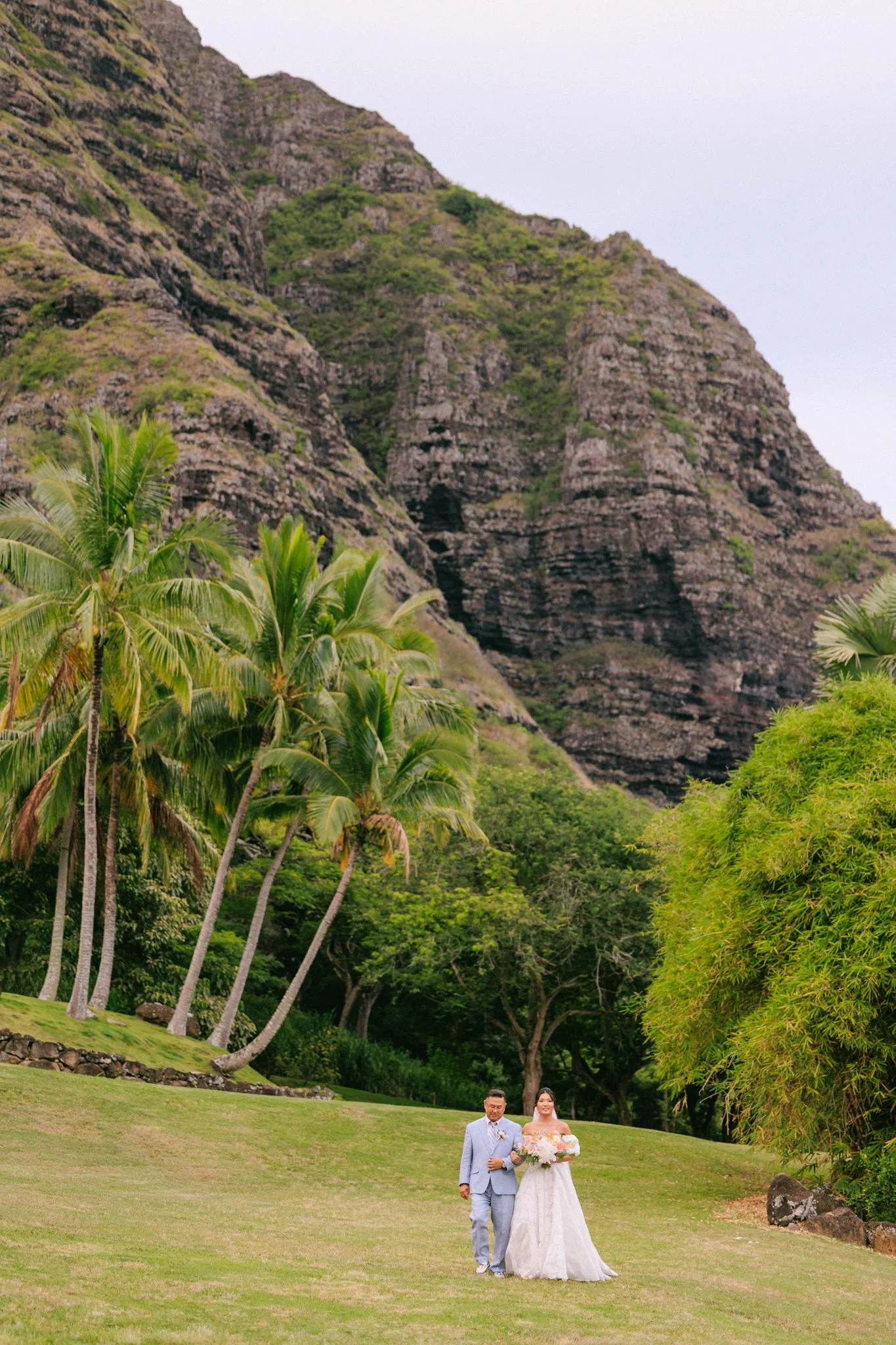 A wedding couple walking on a grassy lawn surrounded by palm trees and lush green trees, with mountains in the background.