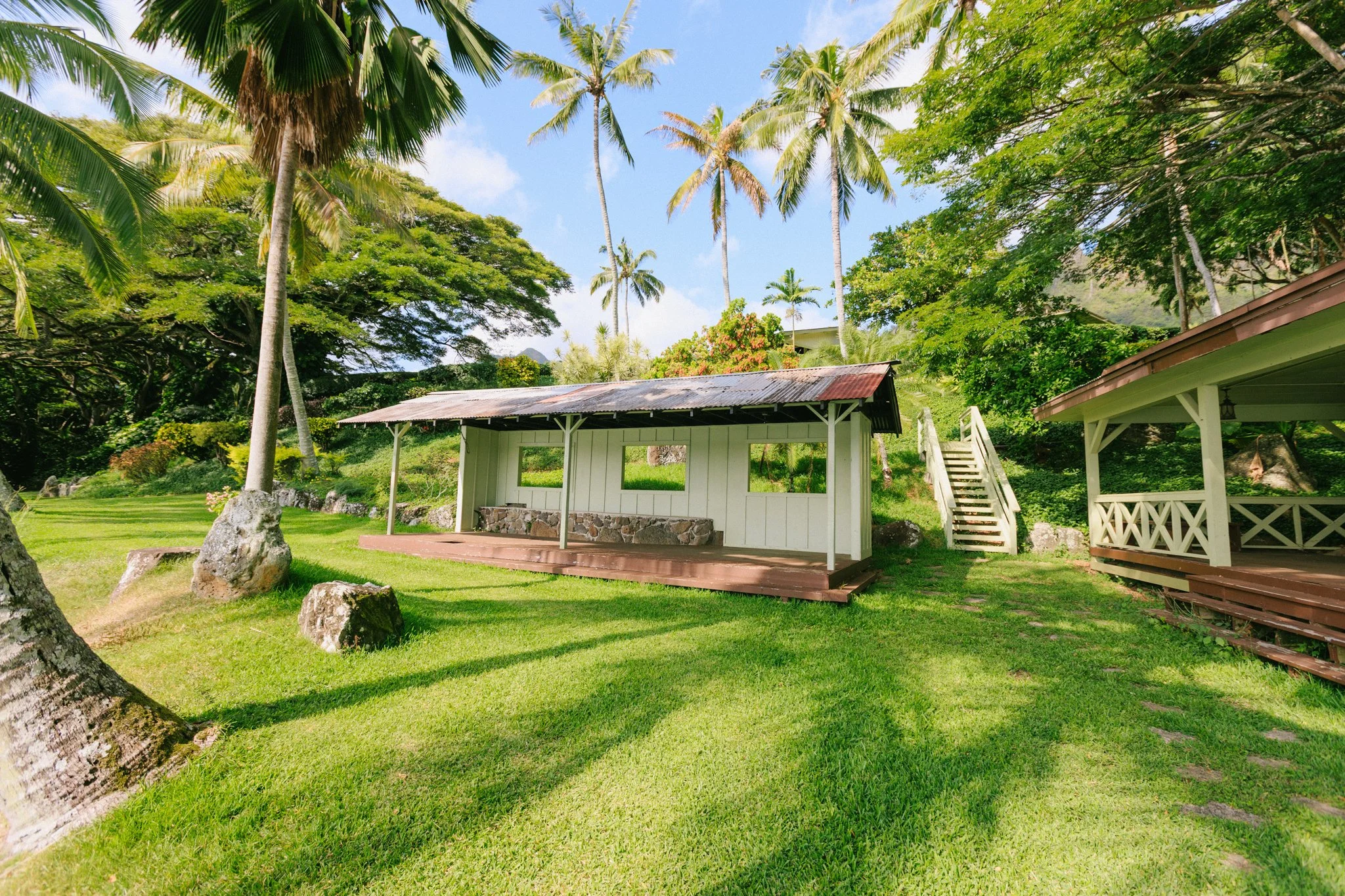 A tropical scene with a small white wooden shelter with a corrugated metal roof, a wooden porch, and stairs leading up to a lush, green hillside. Tall palm trees and other tropical plants surround the area, with a bright blue sky overhead.