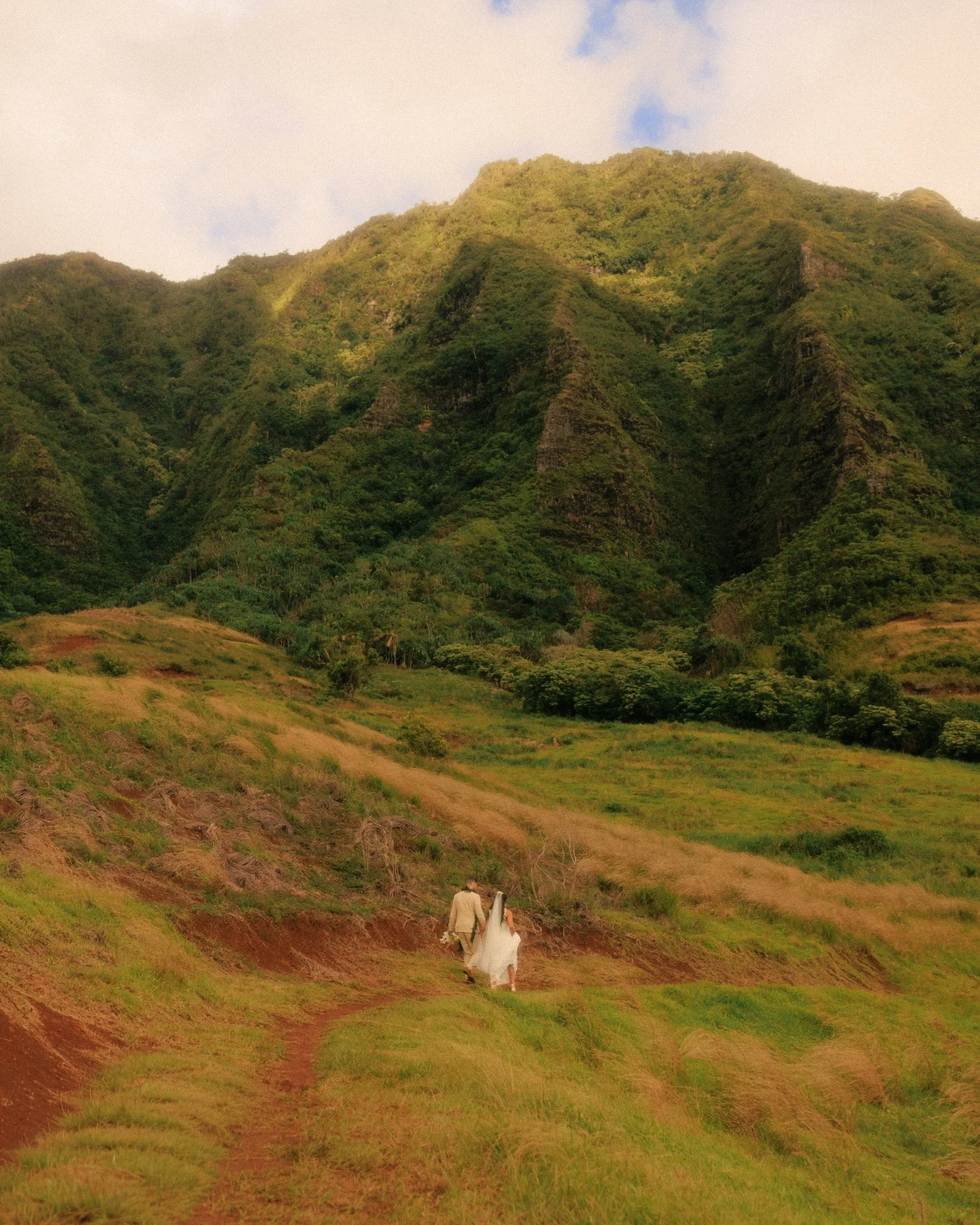 Nicole and Ethan walk small along a dirt path through rolling grass hills beneath a massive green mountain ridge at the Jumanji location in Kualoa Ranch on Oahu, Hawaii, after their Paliku Gardens elopement.