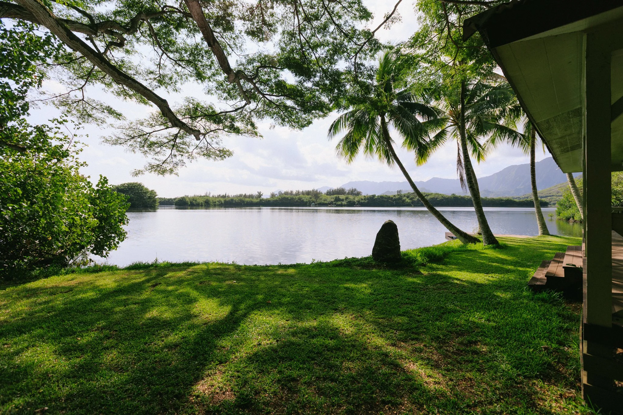 View of a calm lake surrounded by lush green grass, trees, and mountains in the distance, with a house porch on the right side.