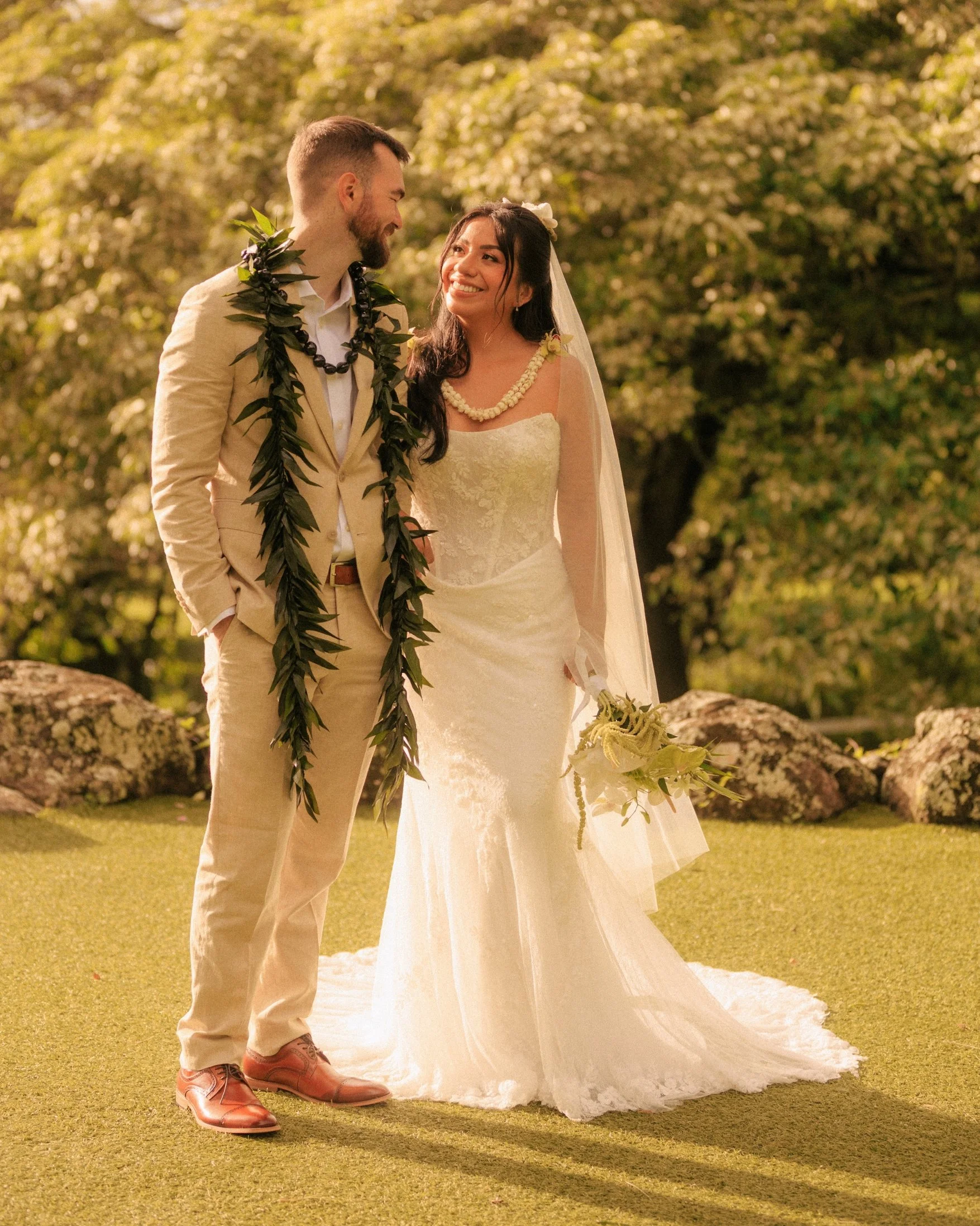Nicole and Ethan stand full-length on the lawn laughing at each other, Ethan wearing a long green leaf lei and Nicole holding greenery, with sunlit trees and rocks behind them at Paliku Gardens, Kualoa Ranch, Oahu.