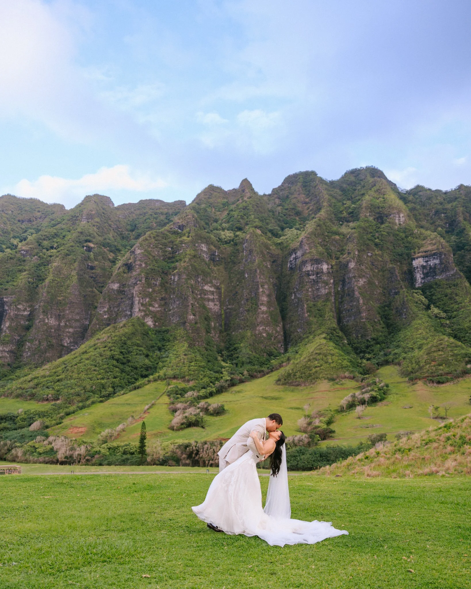 A bride and groom are romantically kissing on a green field with a mountainous landscape in the background.
