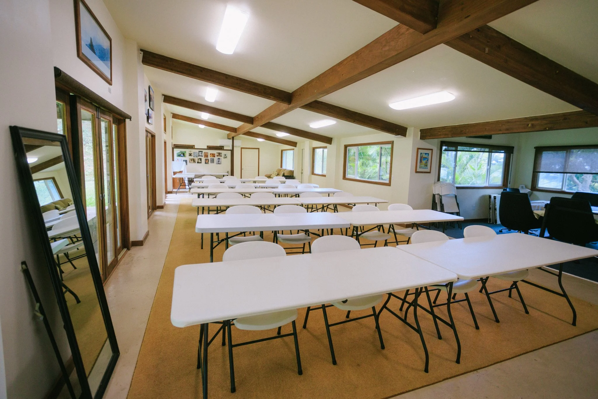 A well-lit conference or meeting room with white rectangular tables and white chairs, large windows letting in natural light, and wood beams on the ceiling. There are also some framed pictures on the walls and a mirror leaning against the wall on the
