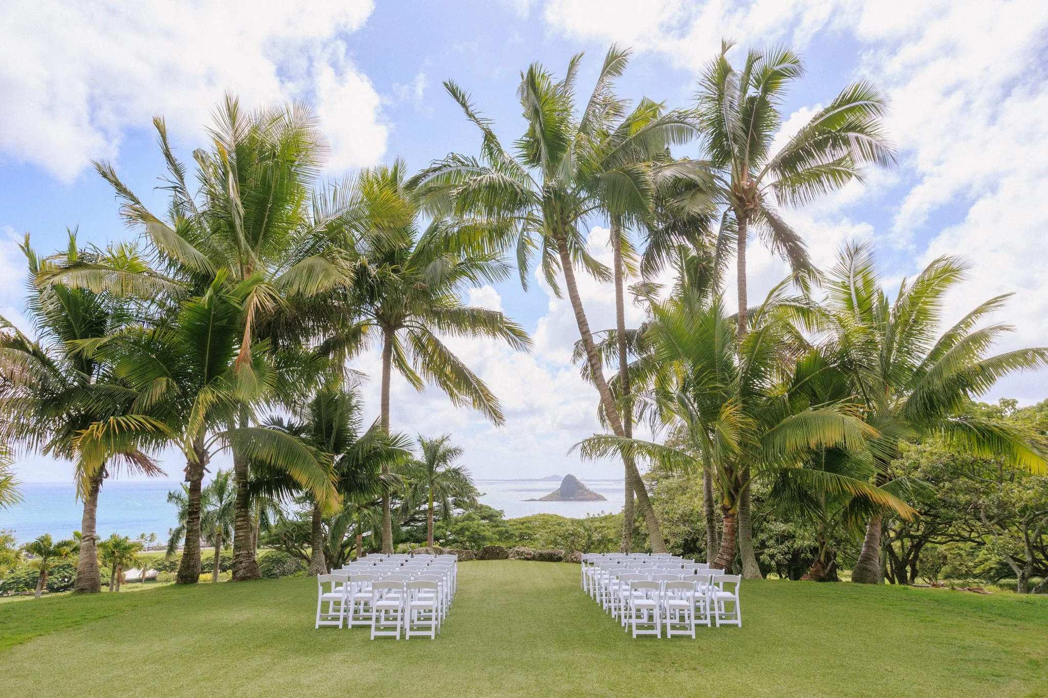 An outdoor wedding setup with white chairs arranged on a grassy lawn, trees, and a view of the ocean and distant islands under a partly cloudy sky.