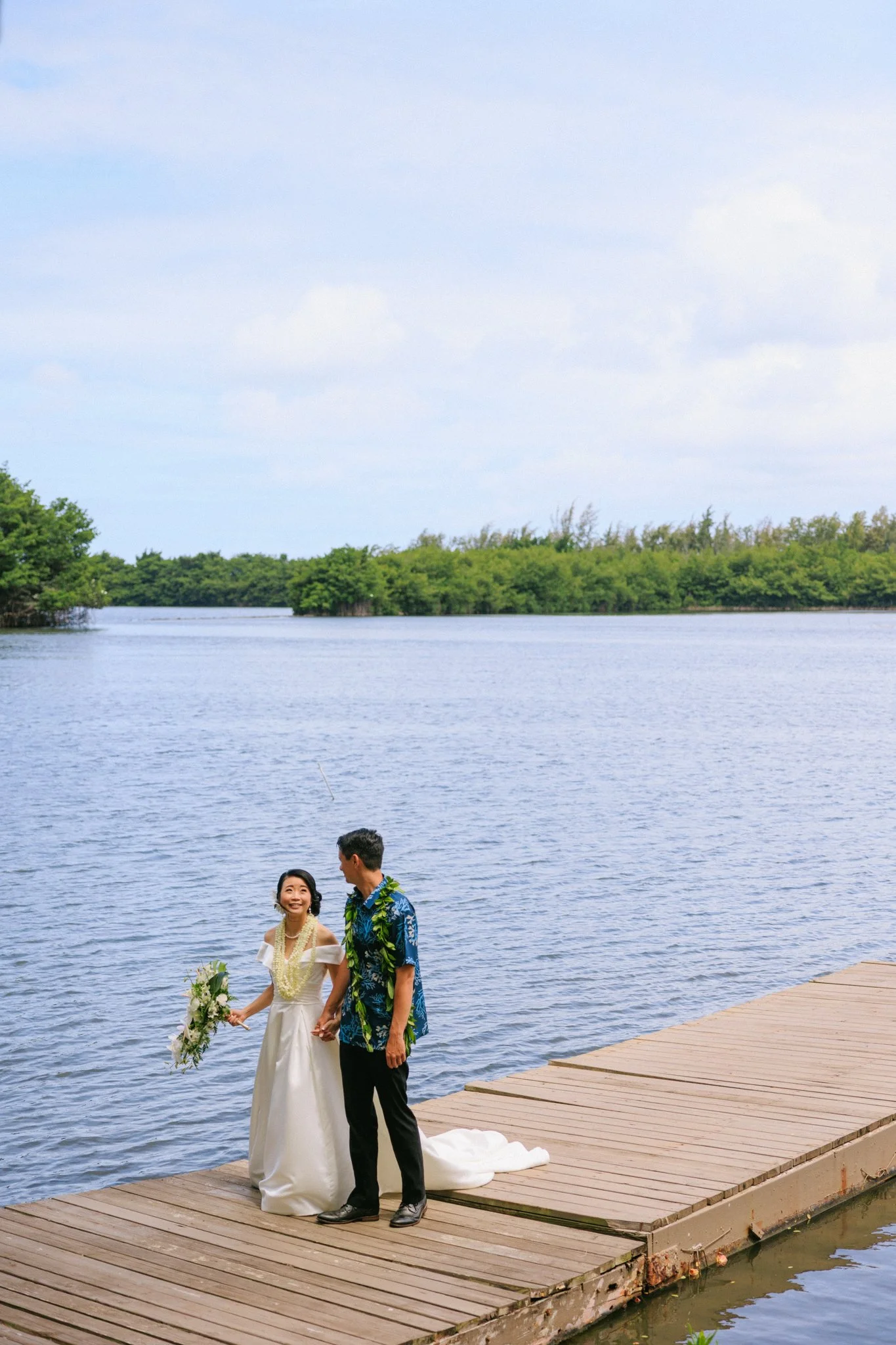 A happy couple holding hands on a wooden dock by a lake, dressed in wedding attire. The woman is in a white wedding dress holding a bouquet, and the man is in a blue Hawaiian shirt and black pants, smiling at each other with a lush green landscape an