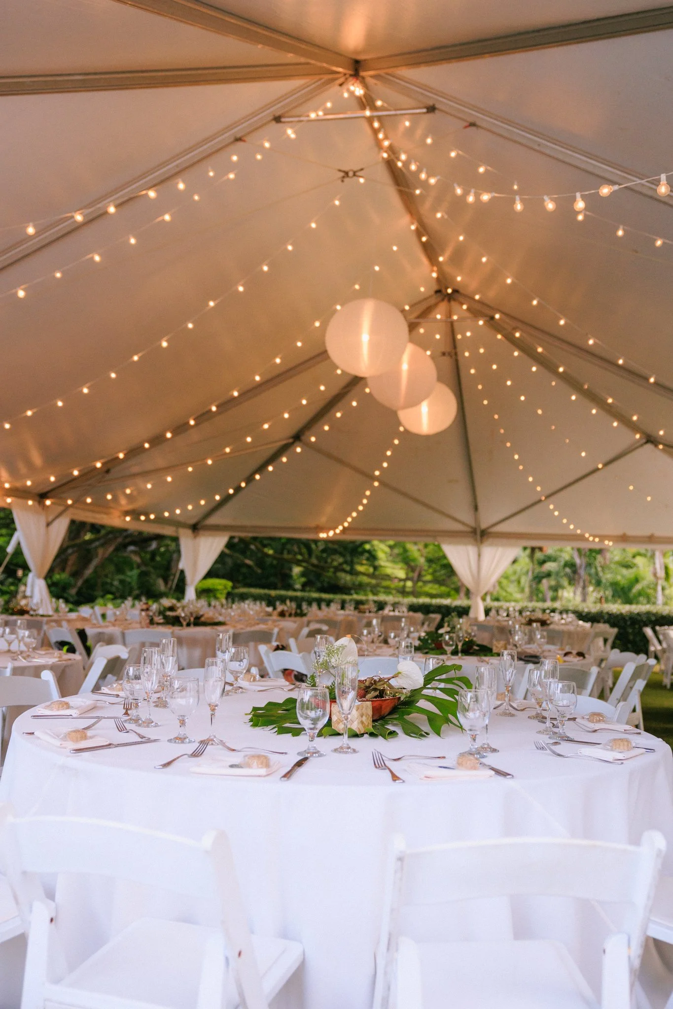 Elegant outdoor event setup under a large tent with string lights and paper lanterns hanging from the ceiling. Round tables with white tablecloths, glassware, and silverware arranged for a formal occasion, with a centerpiece featuring tropical green 