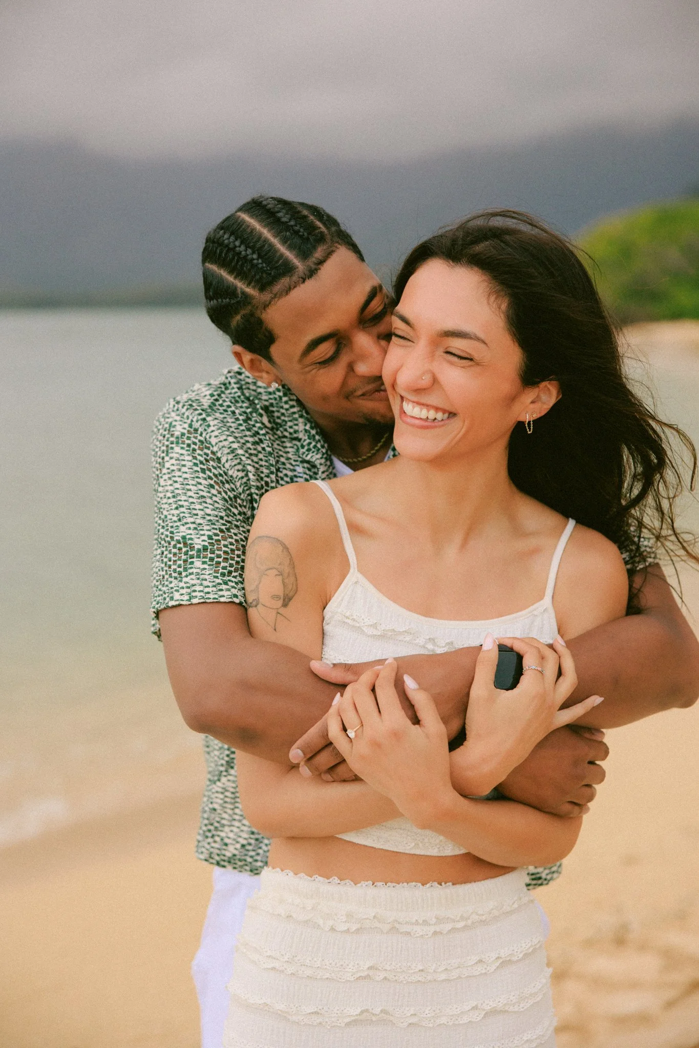 Newly engaged couple embracing on the beach at Kualoa Regional Park after a surprise sunset proposal in Oahu Hawaii.