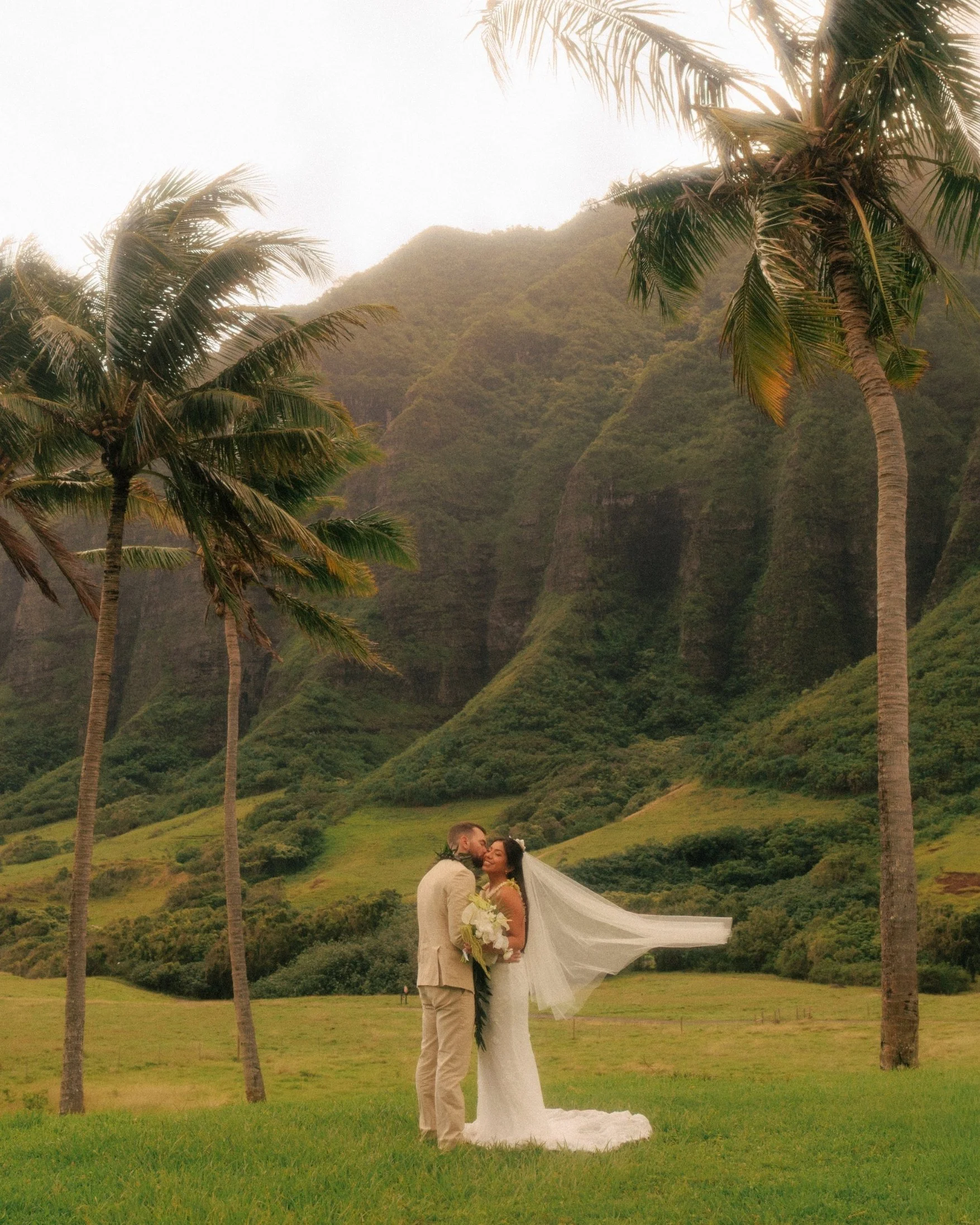 Nicole and Ethan kiss between two tall palm trees with Nicole's veil blowing sideways and the green Ko'olau mountains rising behind them at Kualoa Ranch on Oahu, Hawaii, in a cinematic elopement portrait.