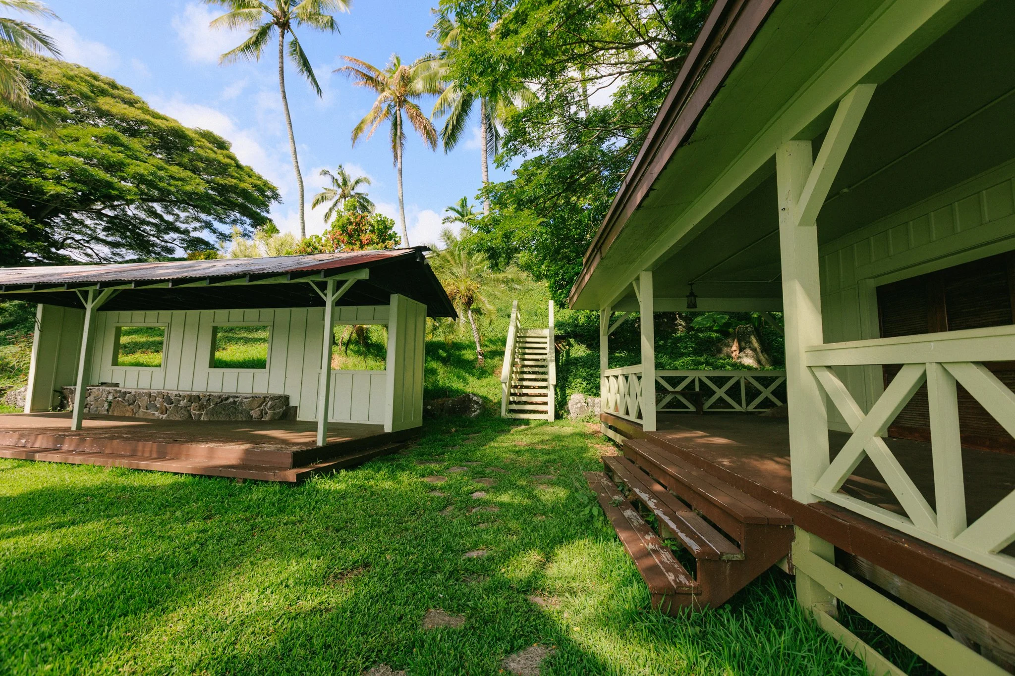 Two green cottages with wooden decks in a tropical setting, surrounded by lush grass, trees, and tall palm trees under a blue sky.